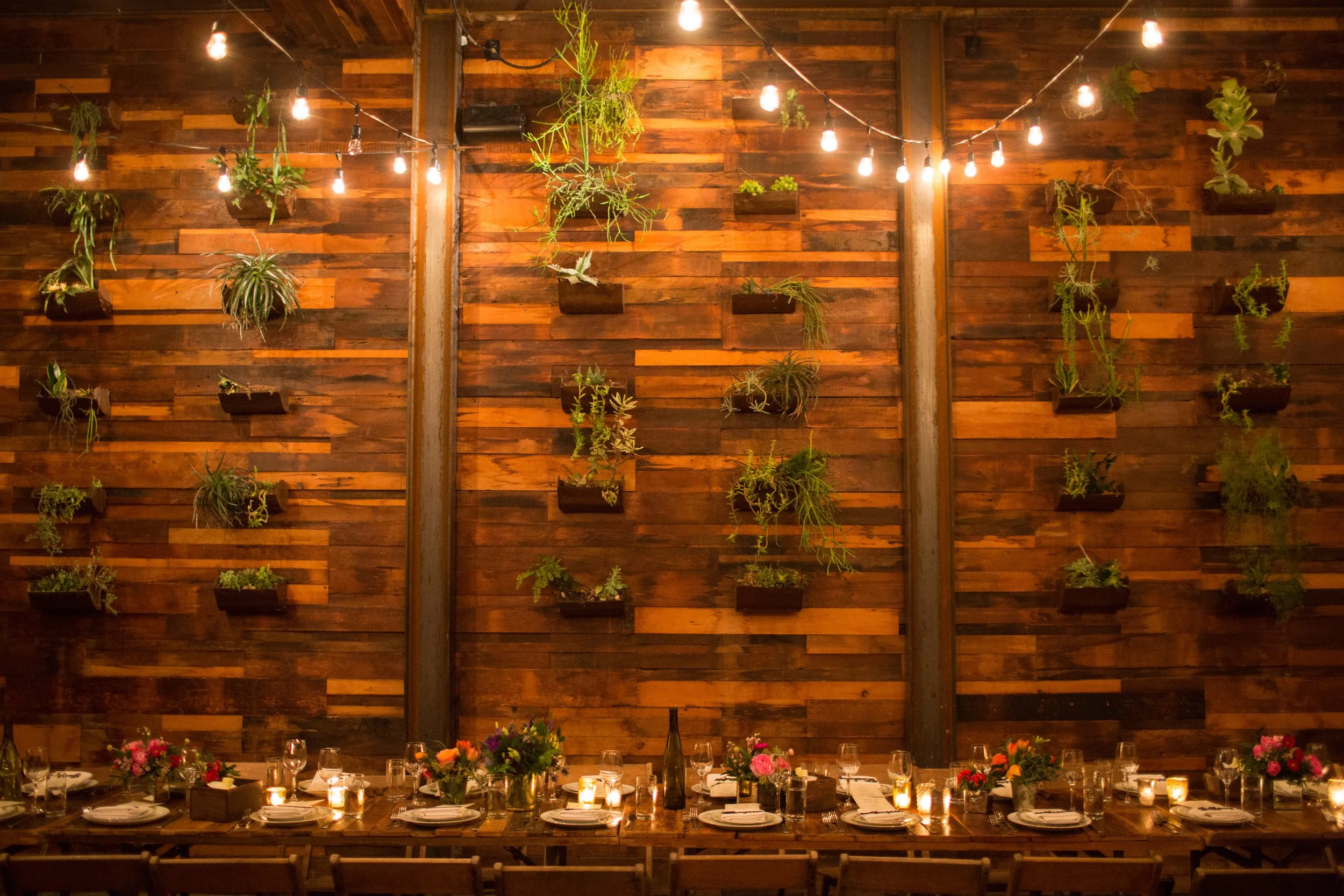 A warmly lit indoor dining area with a long wooden table set with plates, glasses, and candles. The background features a wooden wall decorated with hanging planters and string lights.