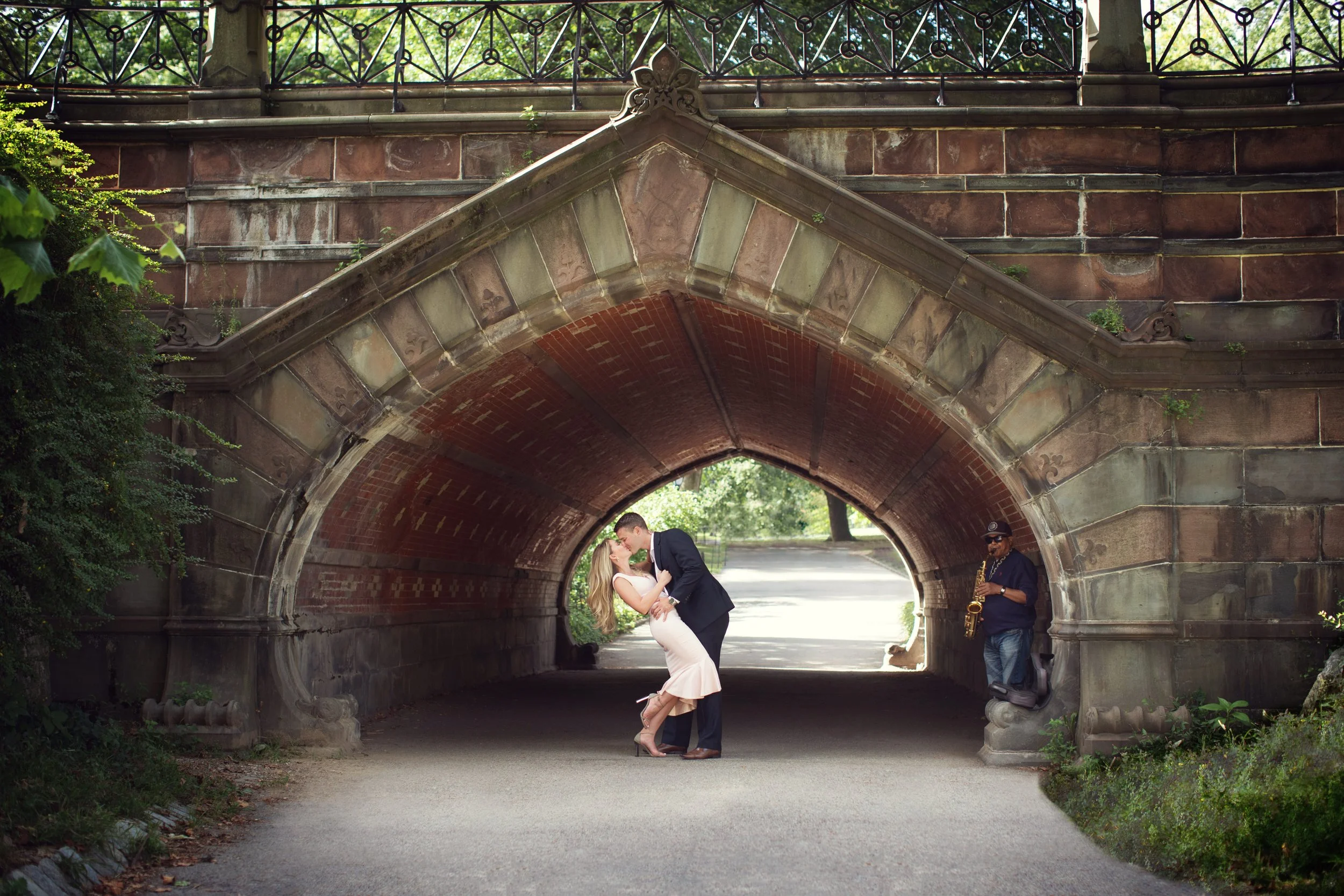 A couple is kissing under a stone bridge, with a saxophone player standing nearby.
