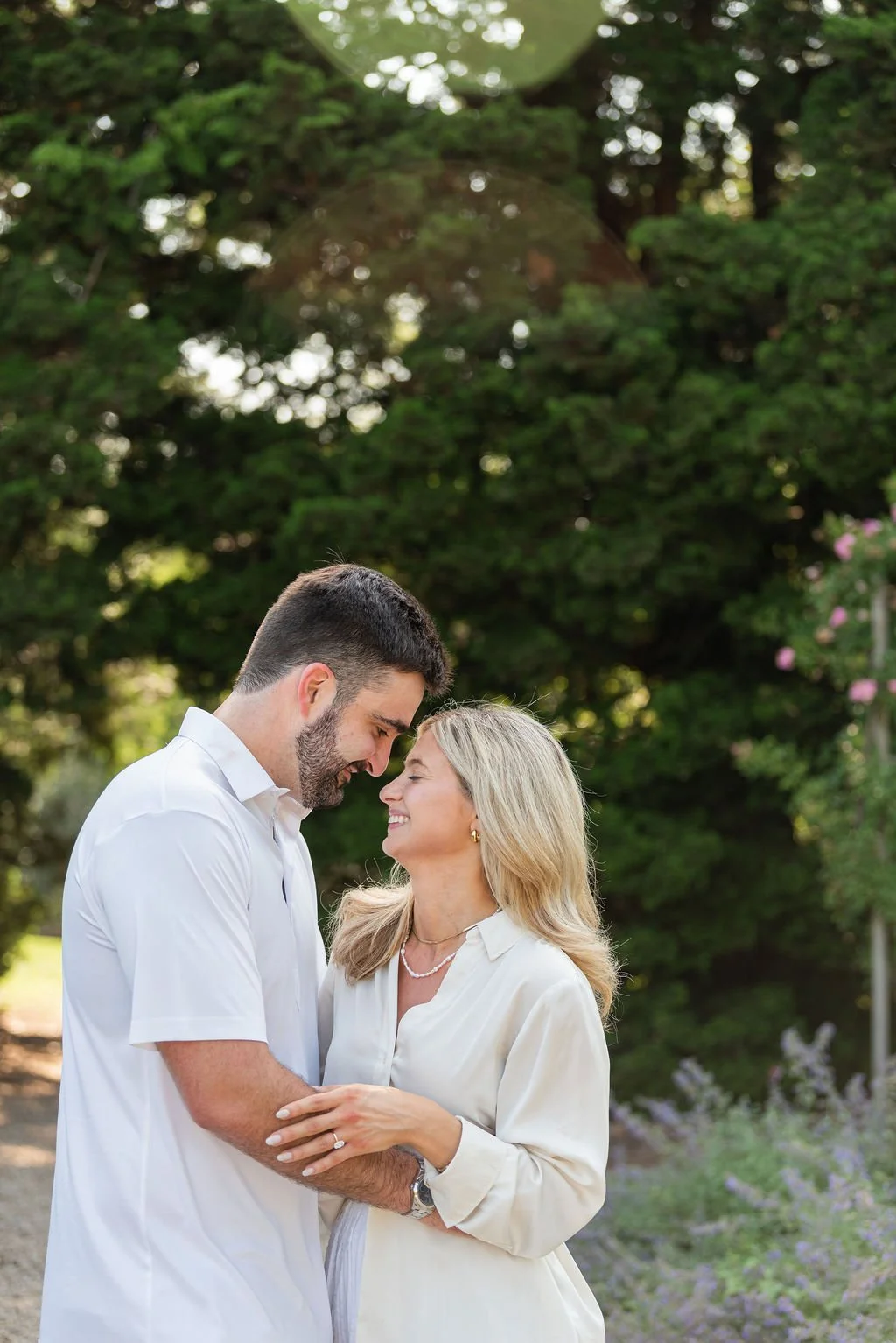 A young man and woman smiling and touching foreheads outdoors during daytime, surrounded by green trees and shrubs.