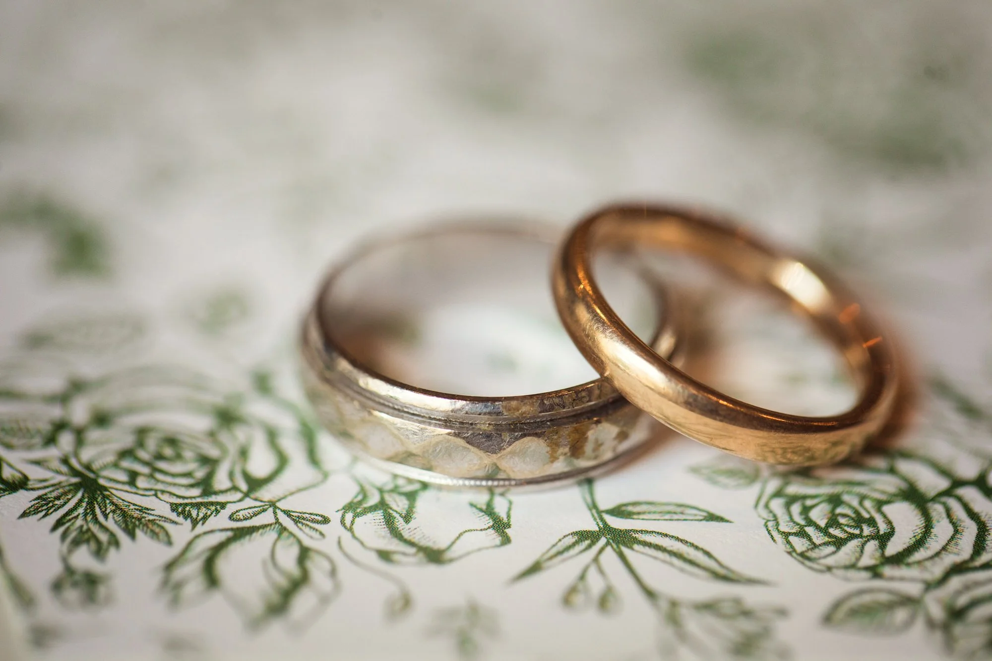 Close-up of two wedding rings, one silver and one gold, placed on a floral patterned surface.