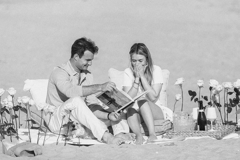 A couple sitting on a bed with pillows, surrounded by white roses and picnic items, with a small dog; the man is showing a book to the woman, who appears surprised or excited.