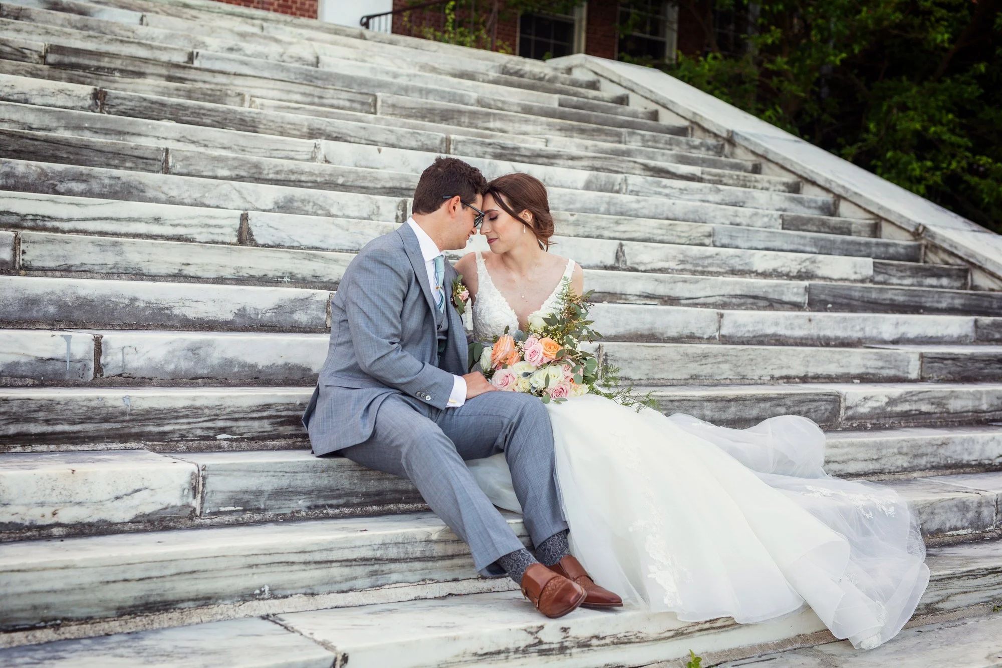 A bride and groom sitting on marble steps, foreheads touching, with the bride holding a bouquet of flowers.