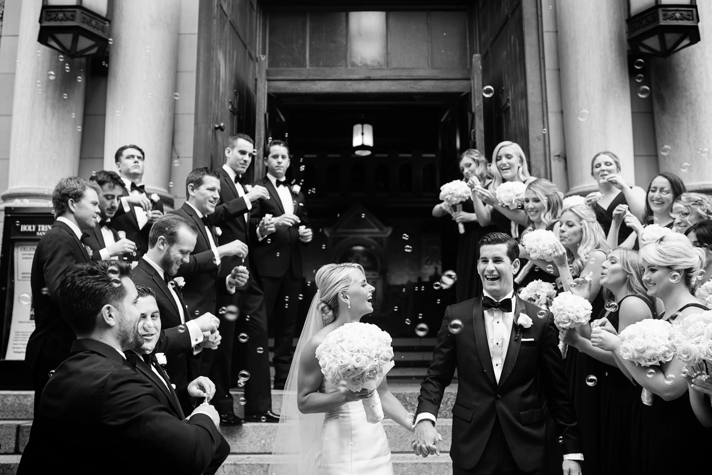 A black and white photo of a wedding celebration outside a church with the bride and groom holding hands in the center, surrounded by bridesmaids and groomsmen blowing bubbles and smiling.