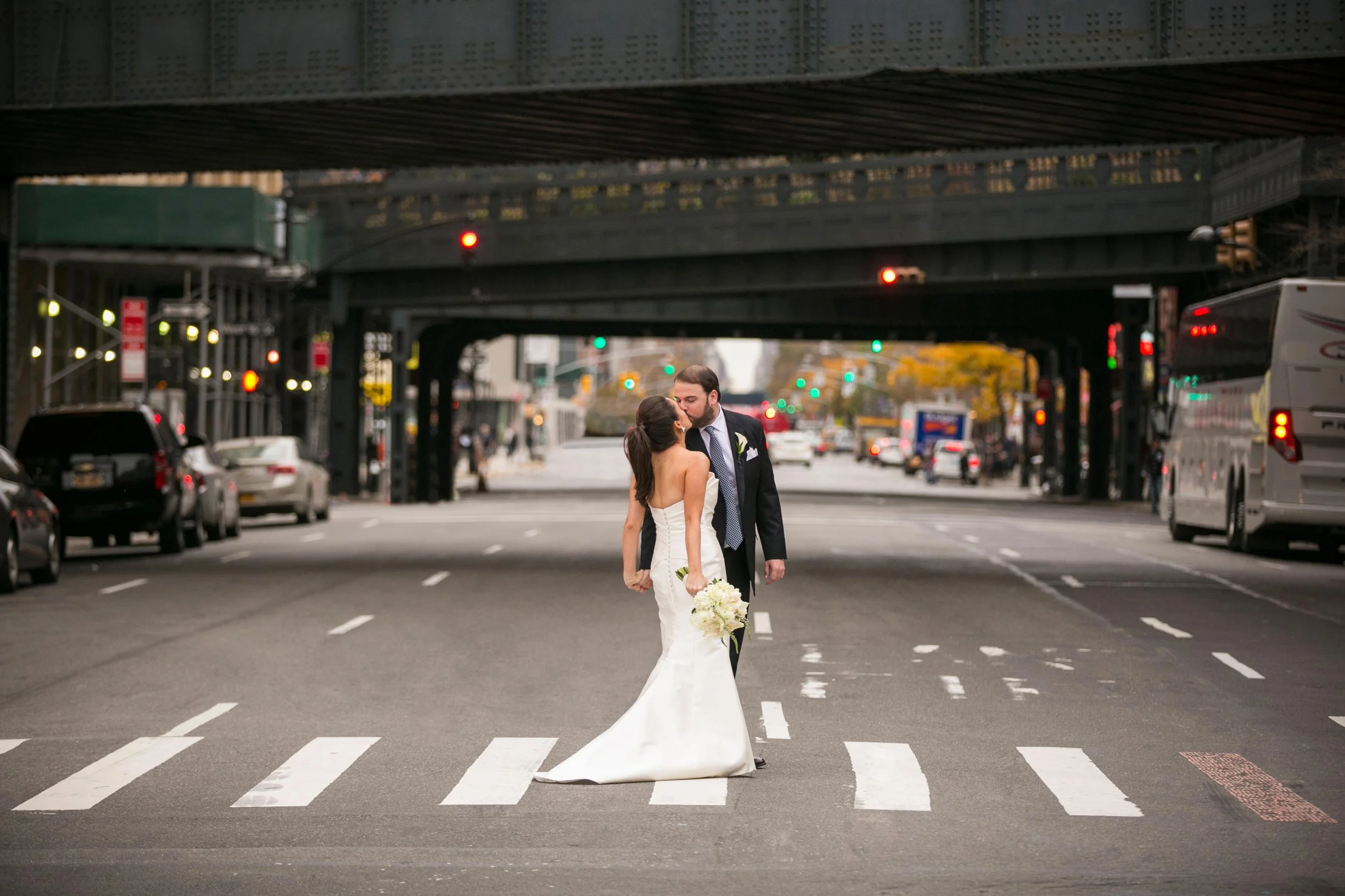 A bride and groom kissing in the middle of a city street under an overpass, with cars and traffic lights in the background.
