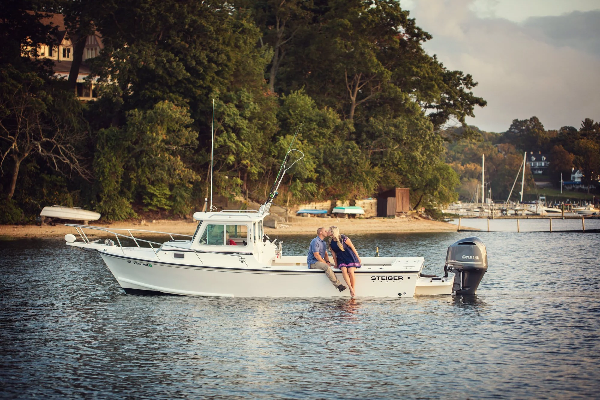 A couple sitting on the edge of a white motorboat in a harbor, kissing with water and trees in the background.
