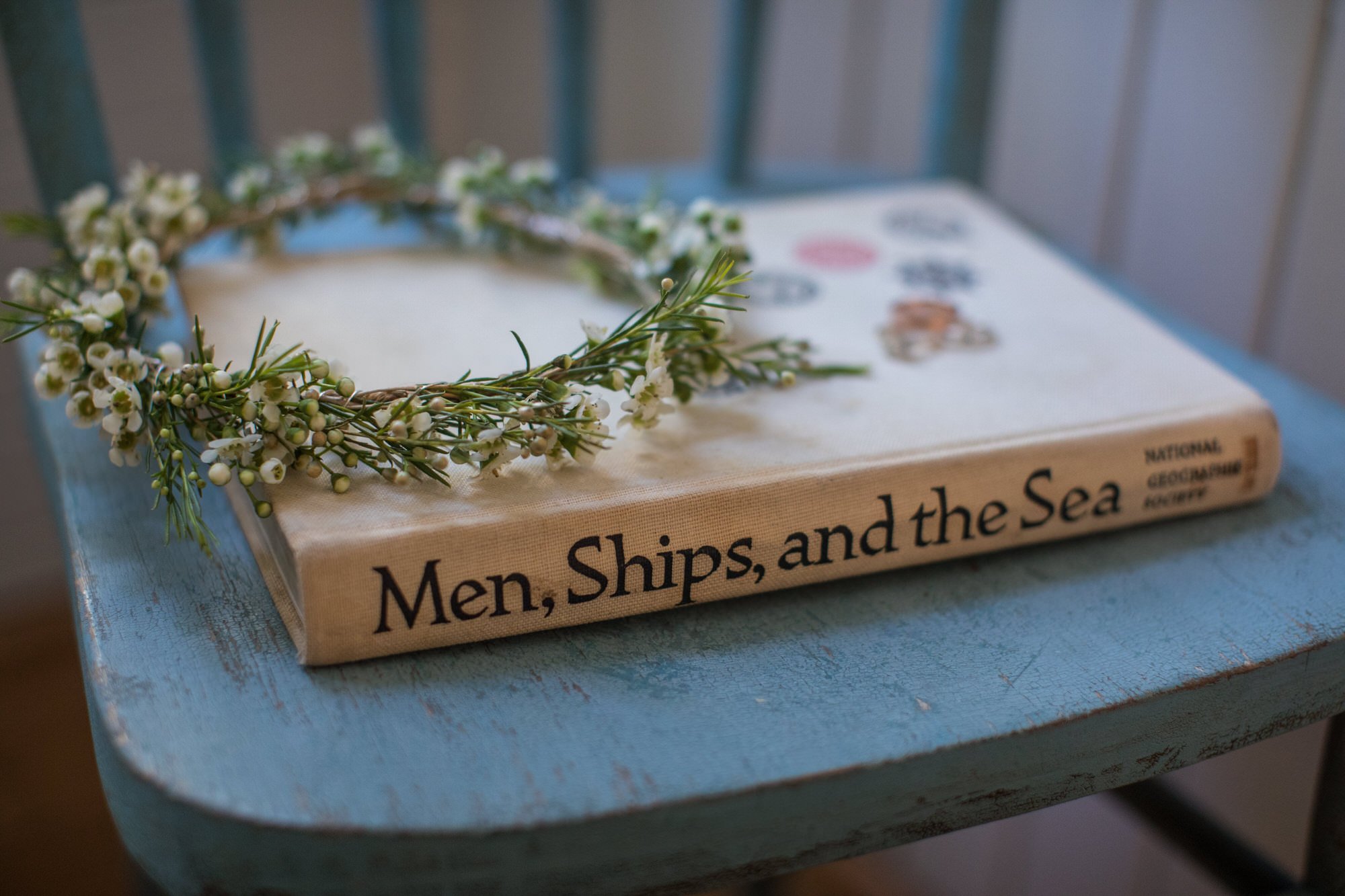 A vintage blue wooden chair with a book titled 'Men, Ships, and the Sea' resting on it. A delicate flower crown made of small white flowers and green leaves decorates the book, placed on a white cloth with printed illustrations and text.