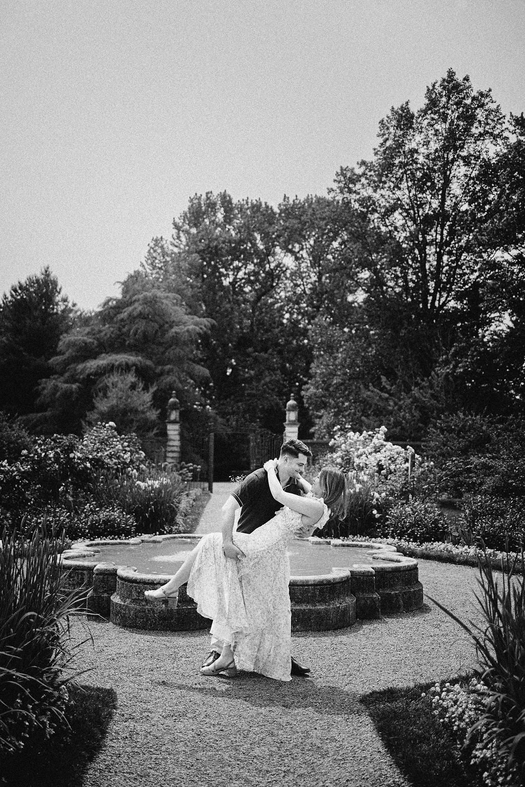 A black and white photo of a couple dancing in a garden with a fountain and trees in the background.