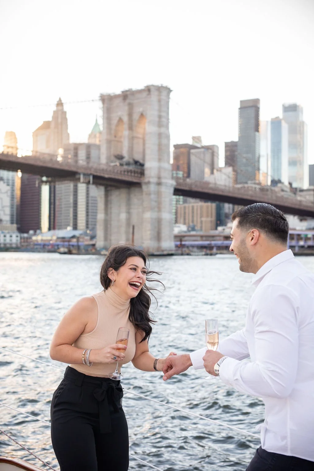 A couple on a boat in New York City, holding glasses of champagne and smiling at each other with the Brooklyn Bridge and Manhattan skyline in the background.