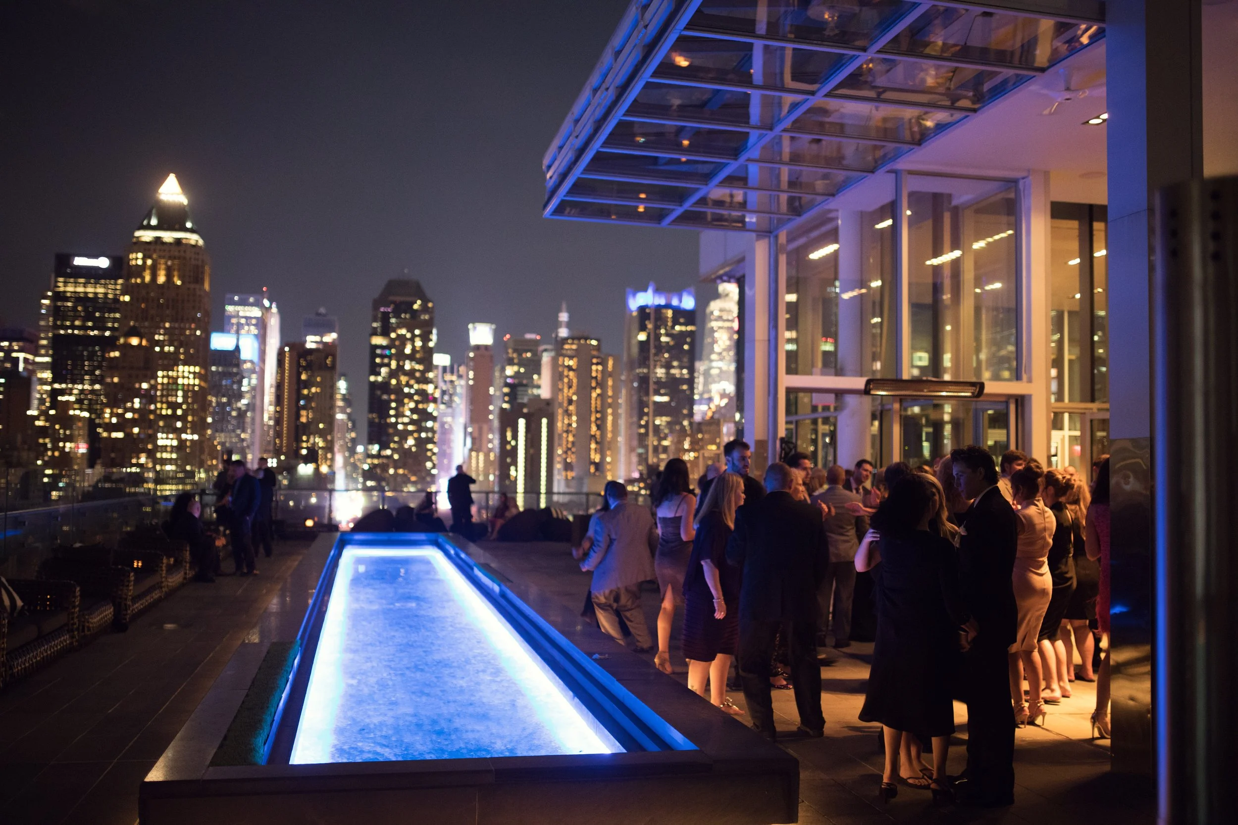 Nighttime rooftop party with city skyline view, illuminated swimming pool, and groups of people socializing outside.