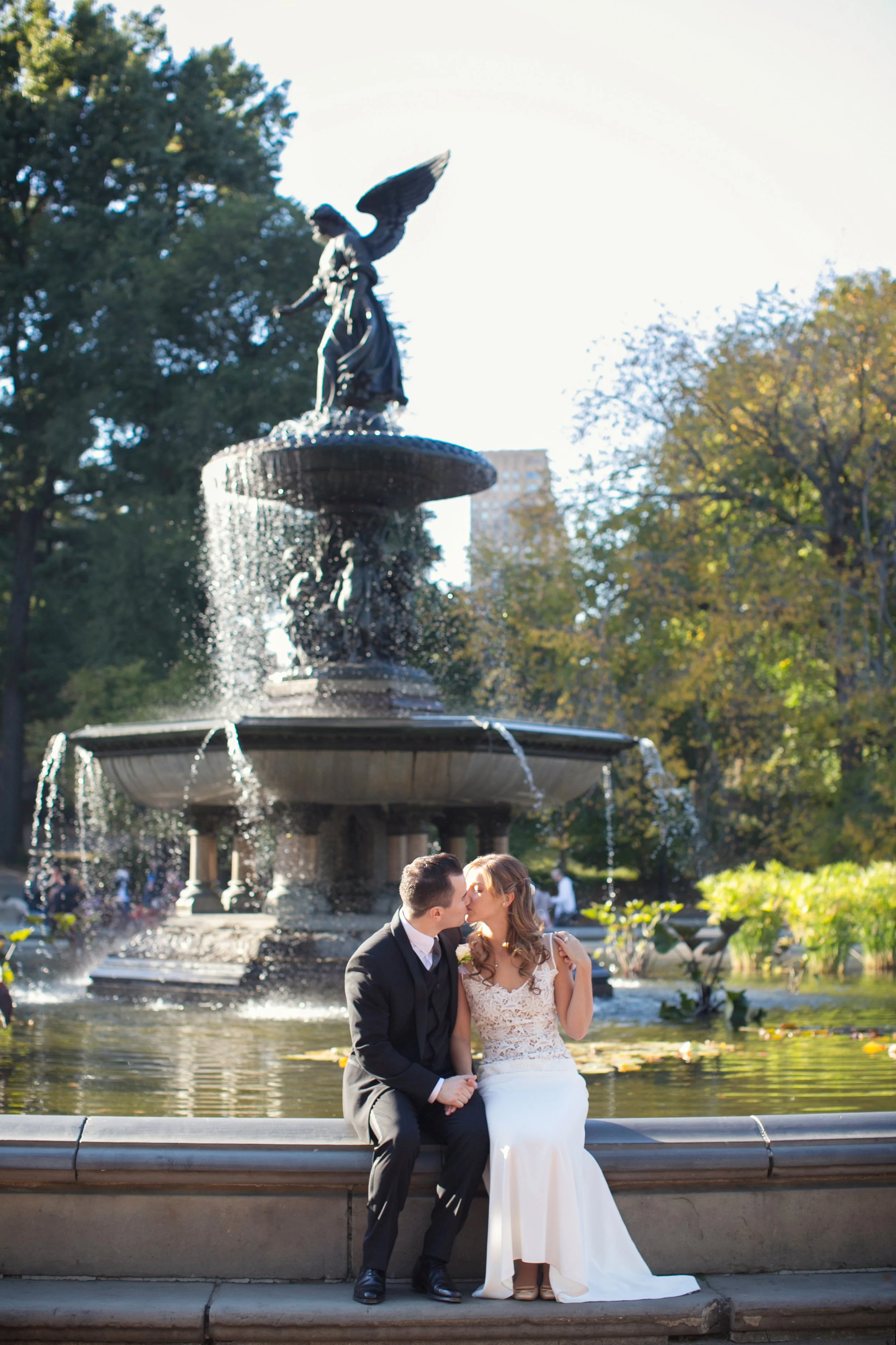 A newlywed couple kisses sitting on the edge of a fountain in a park, with trees and a cityscape in the background.