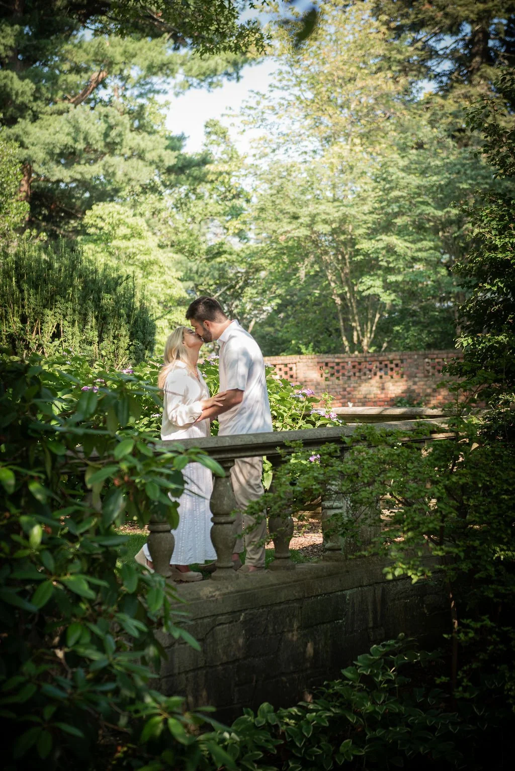 A couple sharing a kiss on a stone bridge in a lush green garden with trees and flowers.