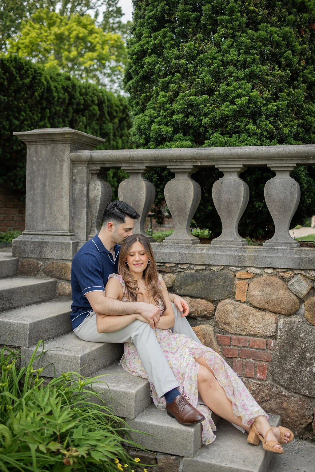 A young man and woman sit on stone steps outdoors, with the man embracing the woman who appears to be upset or crying. Background includes green trees and a stone railing.