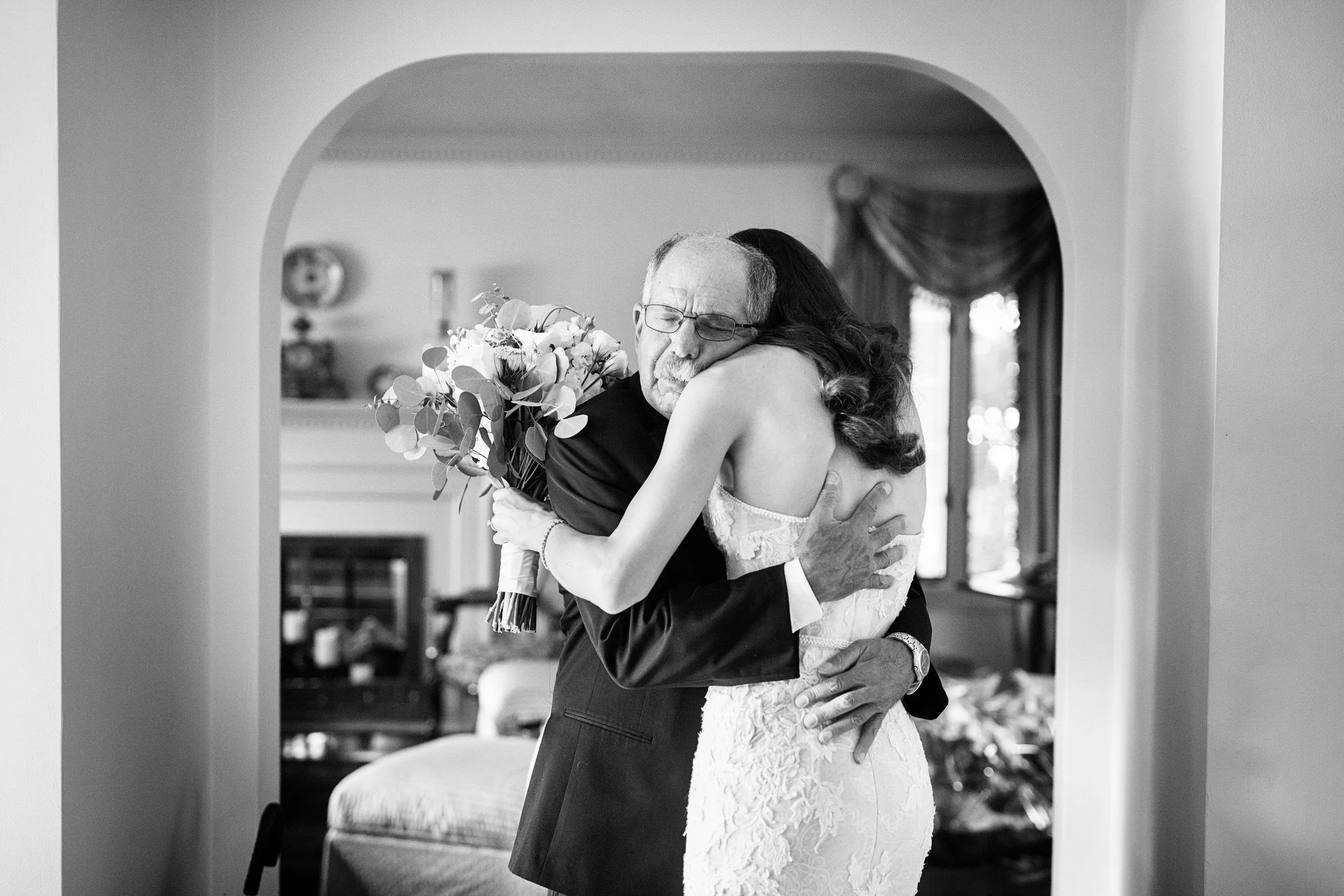 An elderly man and a young woman hugging in a living room, the woman holding a bouquet of flowers, both showing emotion.