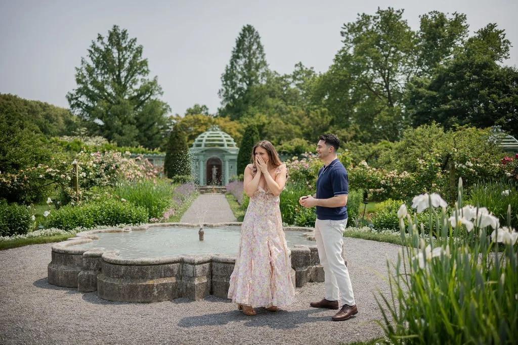 A woman in a pastel floral dress covering her mouth in surprise, standing next to a man in a blue sweater and white pants, smiling in a garden with a fountain and lush greenery.