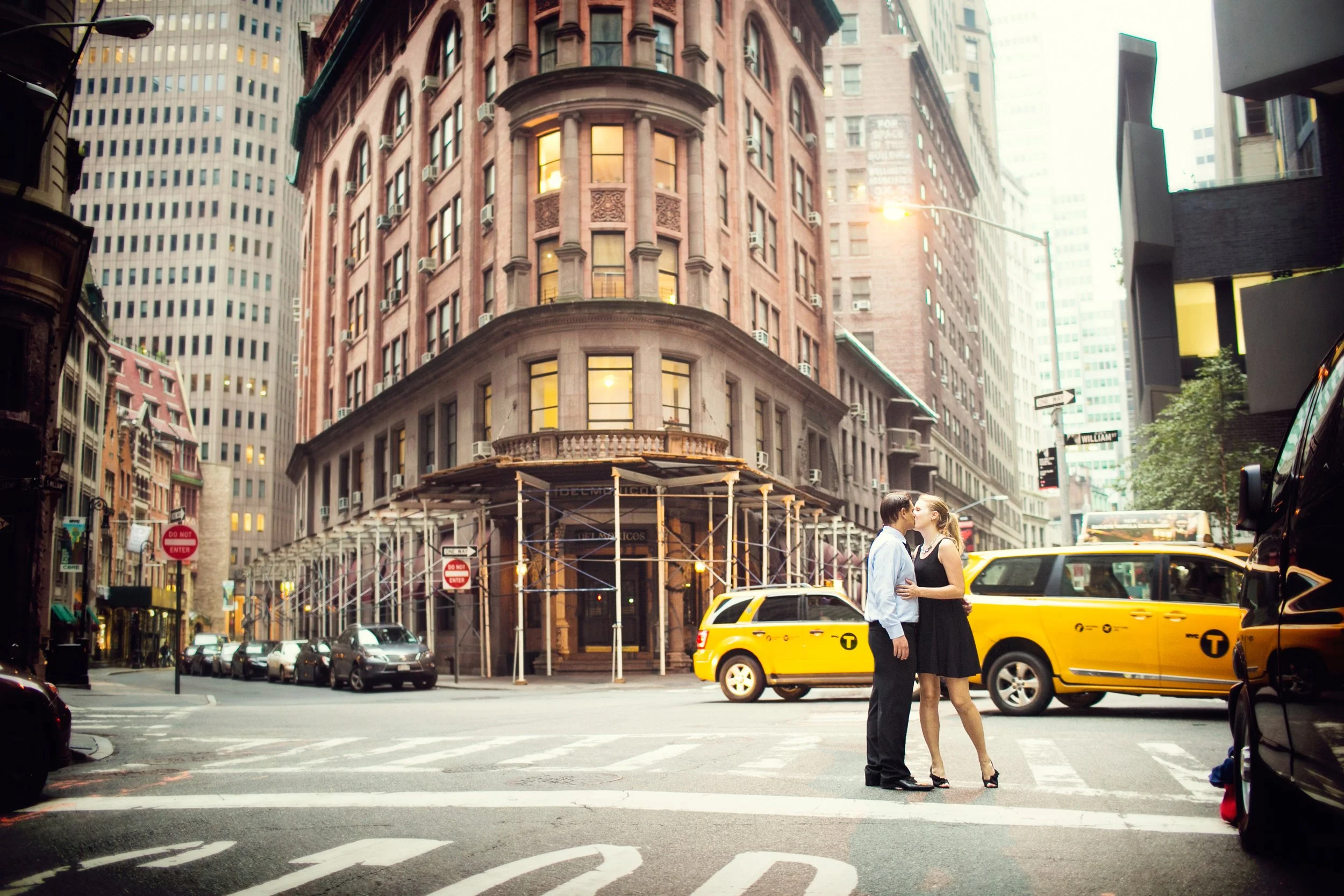 A couple kissing in the middle of a city street with yellow taxis, tall buildings, and scaffolding in the background.