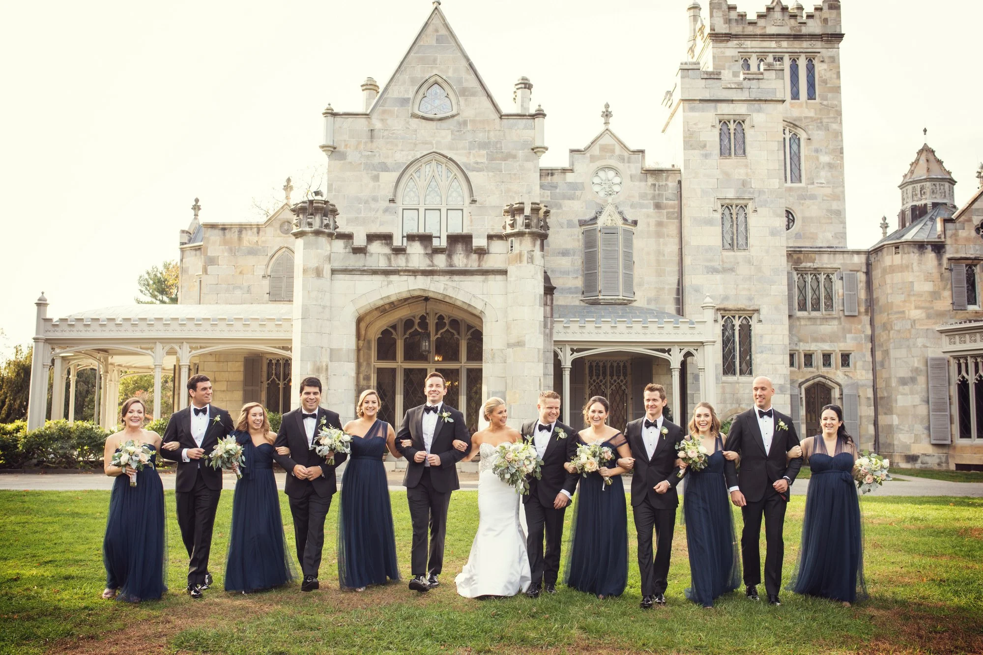 A wedding party posing outside a large stone castle, including the bride, groom, bridesmaids, and groomsmen, all dressed formally and smiling.