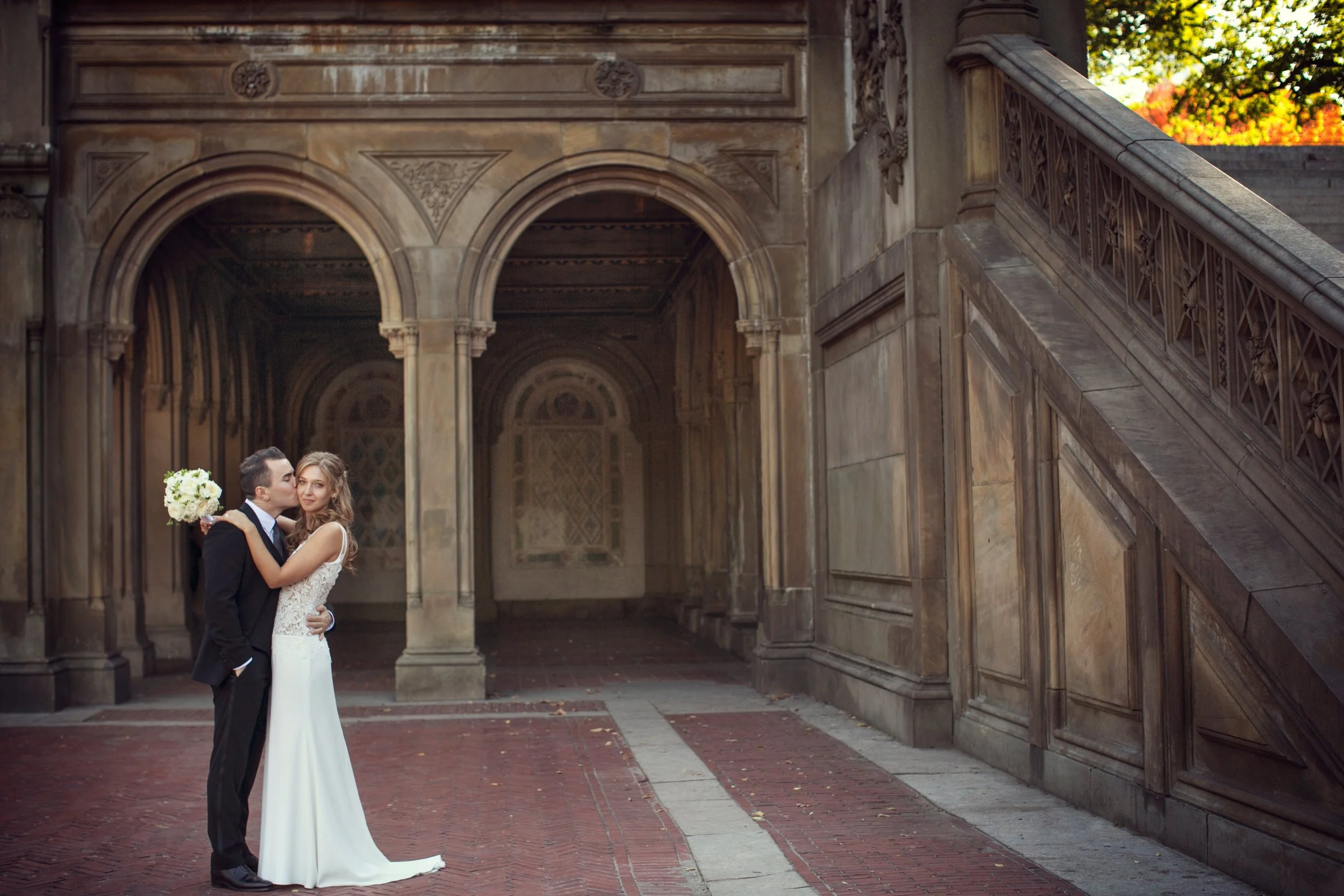 A bride and groom embrace in front of an ornate, historic building with arches and detailed stonework during their wedding.