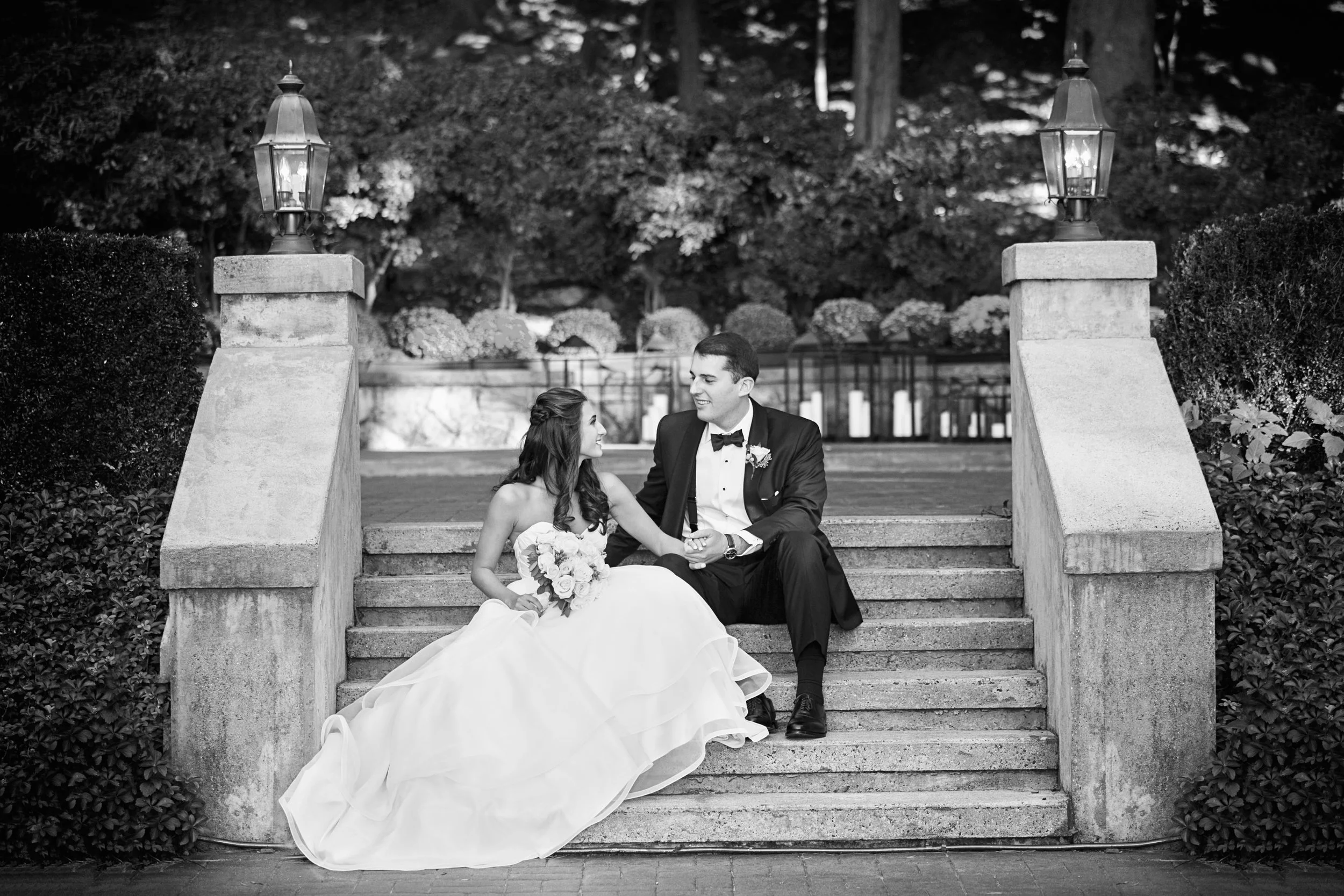 Black and white photo of a bride and groom sitting on outdoor steps, holding hands, in a garden setting with trees and flowers in the background.