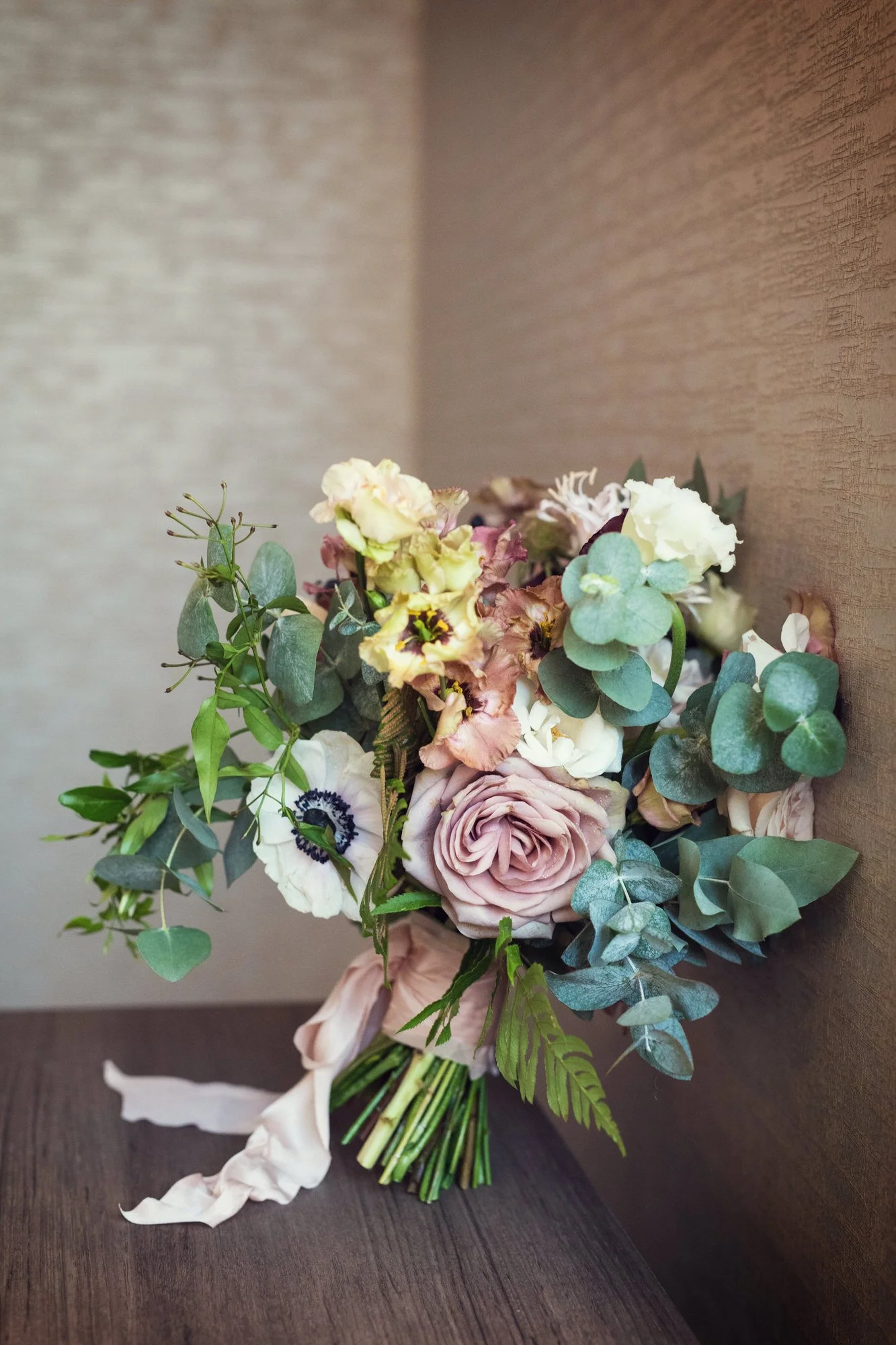 A bouquet of assorted flowers, including roses, anemones, and greenery, tied with a pink ribbon, resting on a dark wooden surface against a textured beige wall.