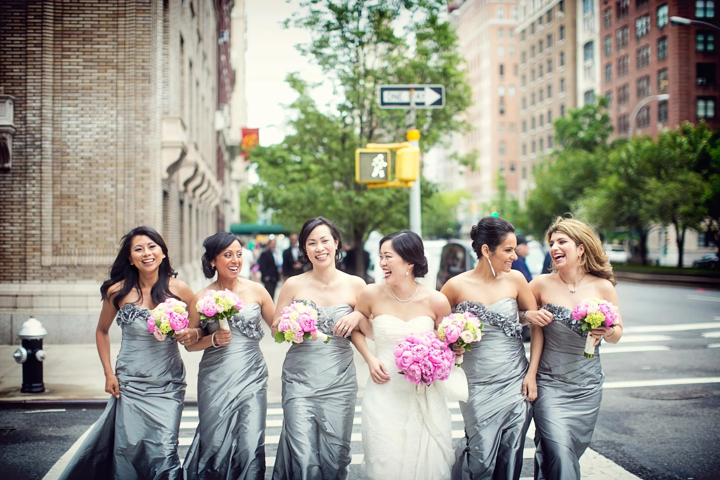 A bride in a wedding dress walking with five bridesmaids in gray dresses. They are smiling and holding bouquets of pink and white flowers, walking across a city street.