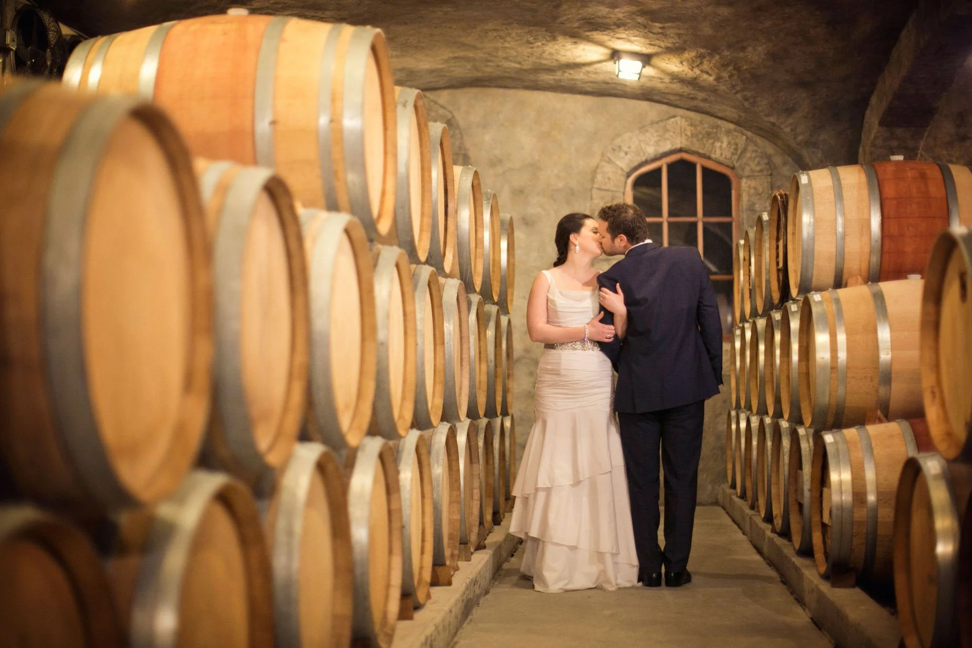 A bride and groom kissing in a wine cellar with wooden barrels stacked on both sides.