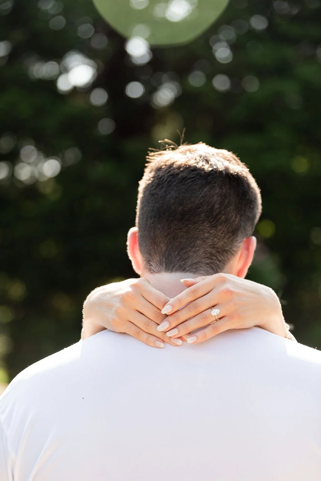 A couple embracing outdoors, with the woman resting her head on the man's shoulder, showing her engagement ring.