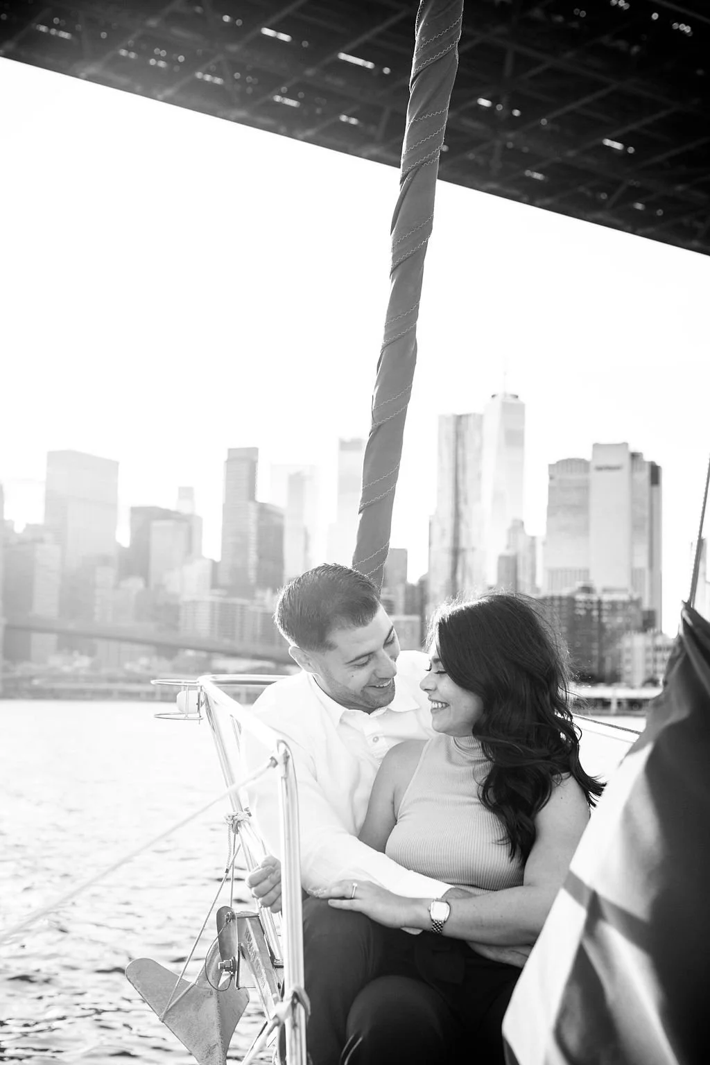 A couple sitting close together on a boat, smiling at each other, with city skyscrapers in the background, in black and white.