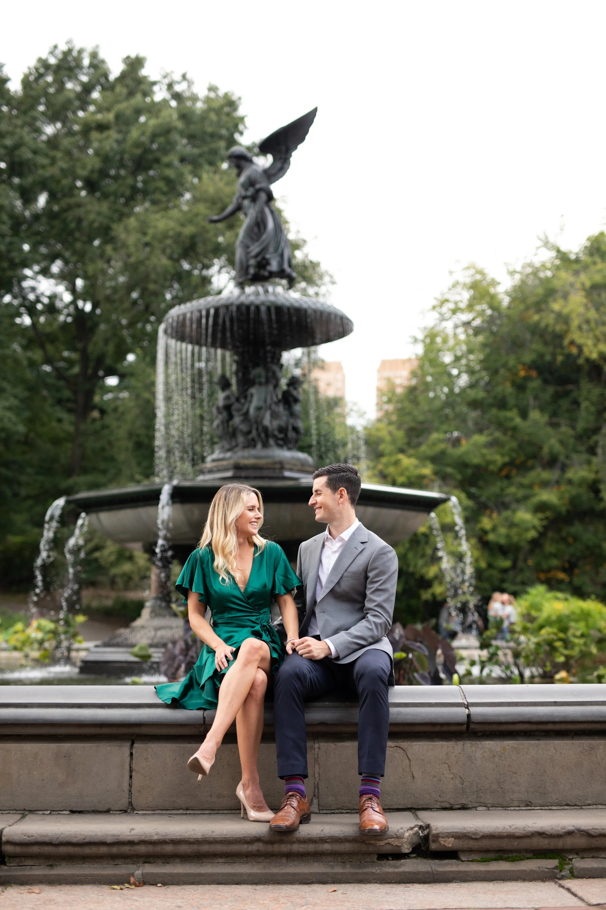 A couple sitting on a fountain bench in a park, smiling and looking at each other, with trees and a statue in the background.
