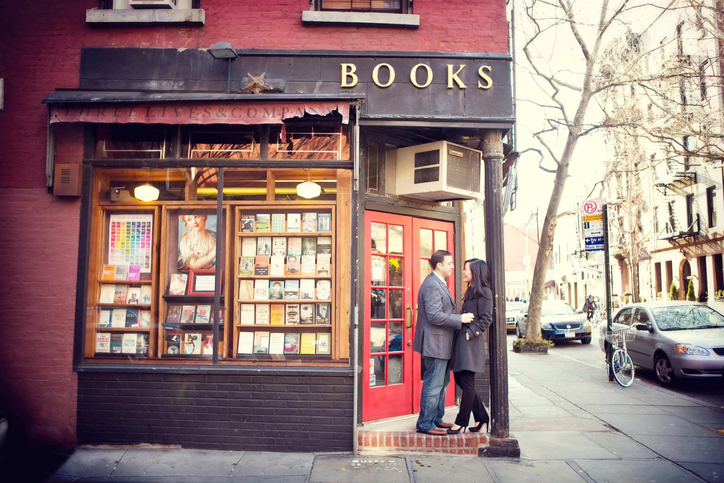 A bookstore storefront with a large window displaying books outside a red door, with three people talking outside on the sidewalk, and a street scene with parked cars and trees in the background.