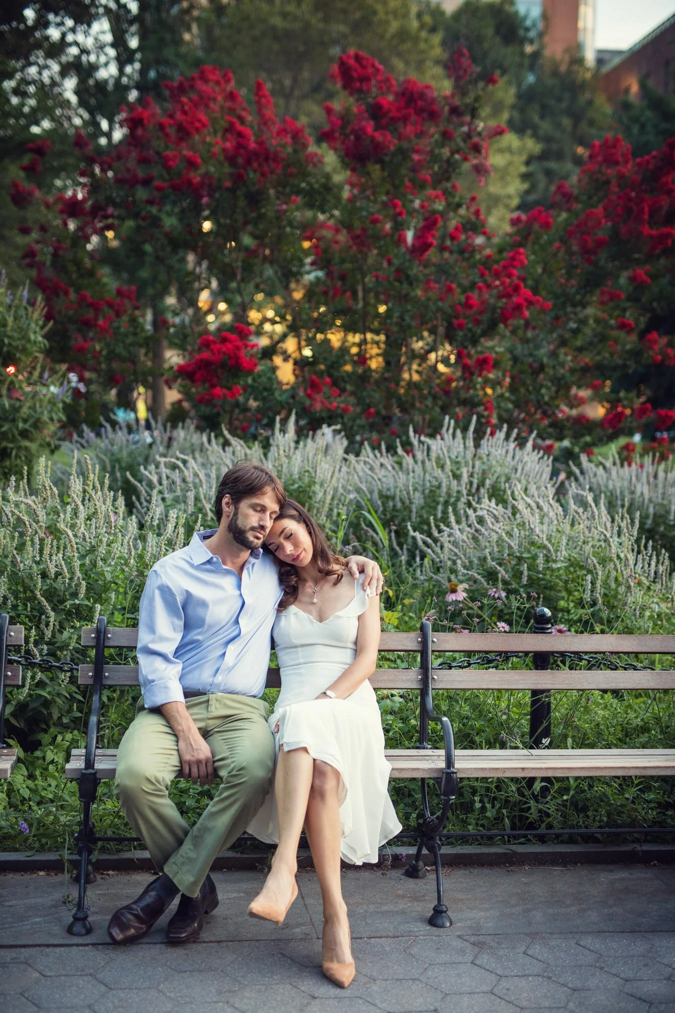 A man and woman sitting closely on a park bench, with their eyes closed, surrounded by colorful flowers and trees.