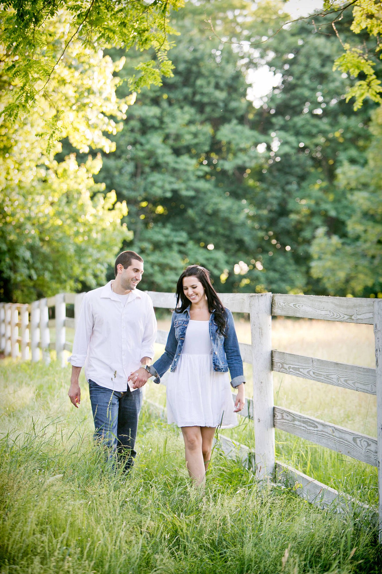 A smiling couple walking hand in hand through a lush green field beside a wooden fence in a park or countryside, with trees and sunlight in the background.