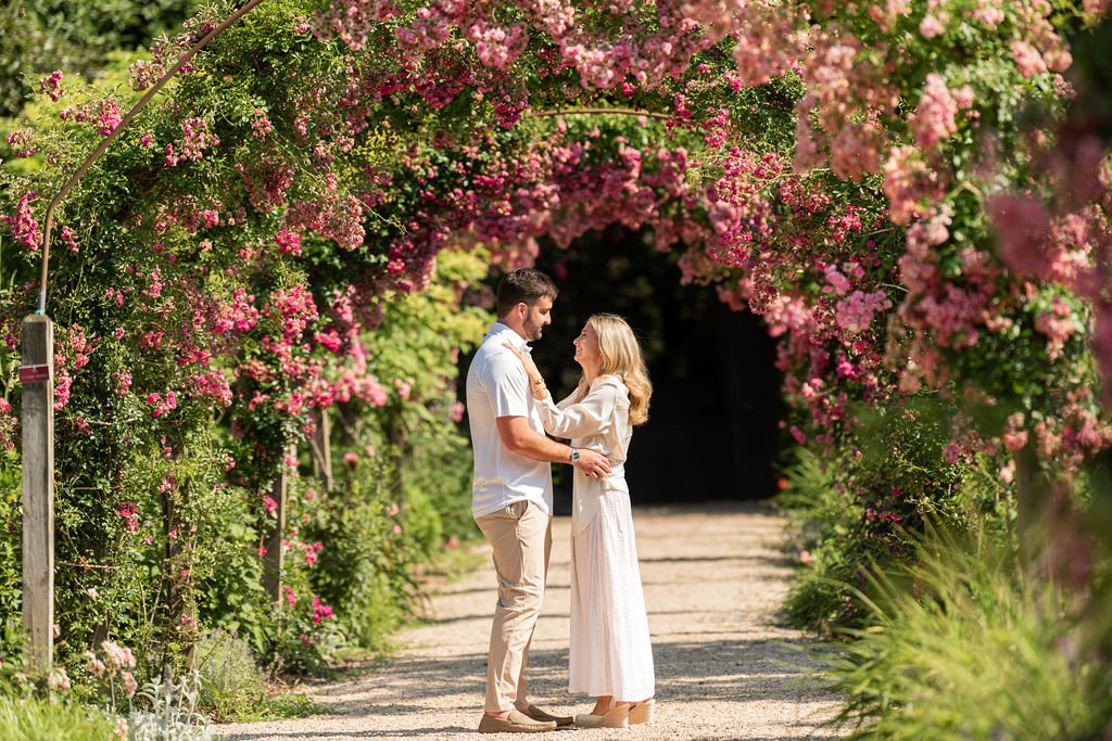 A couple stands in a garden archway covered with blooming pink and purple flowers, sharing a romantic moment.