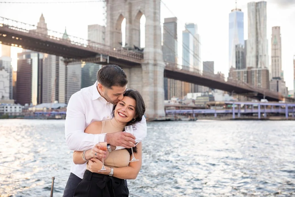A couple enjoying a romantic moment with glasses of champagne by the river, with the Brooklyn Bridge and Manhattan skyline in the background.