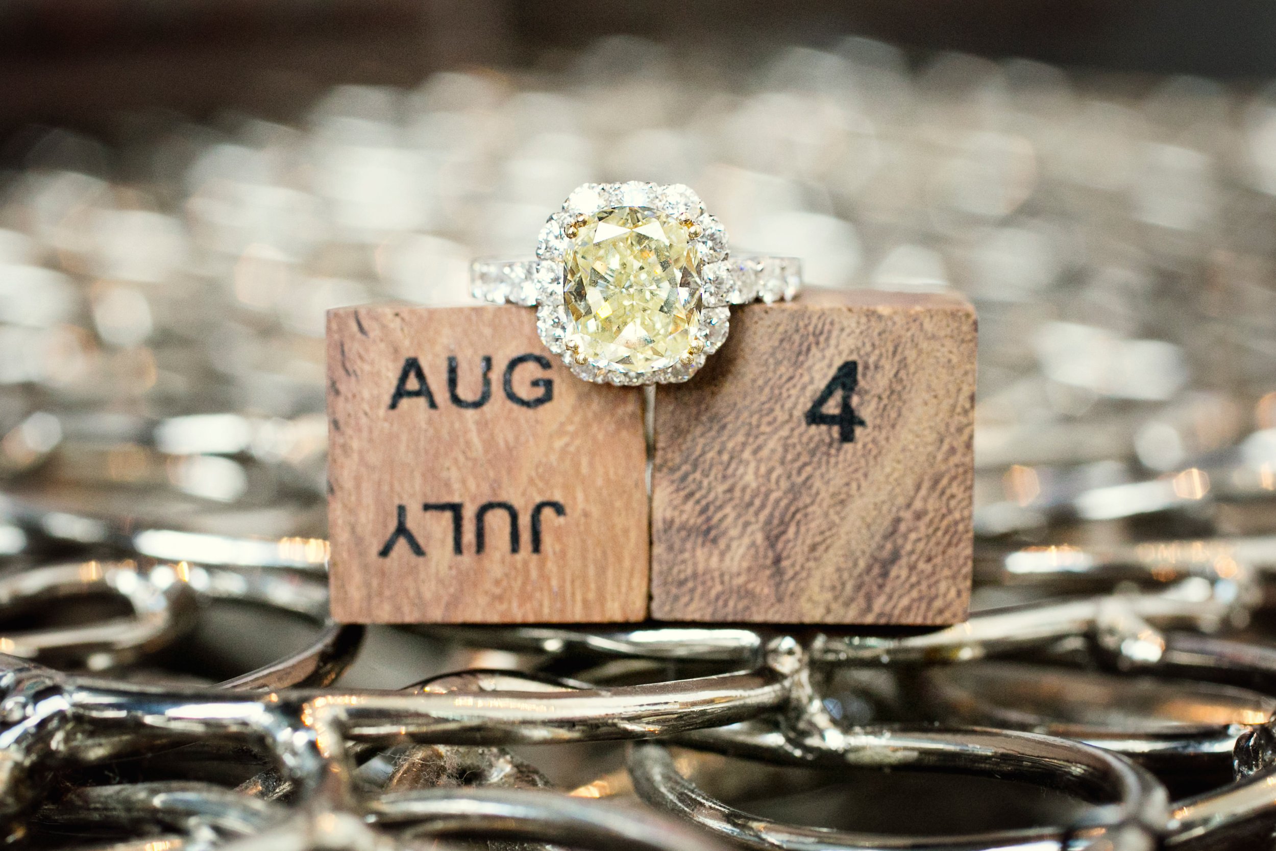 An engagement ring with a yellow diamond surrounded by smaller diamonds is placed on a wooden block with the date August 4, displaying both 'AUG' and '4'. The block is atop a silver chain.