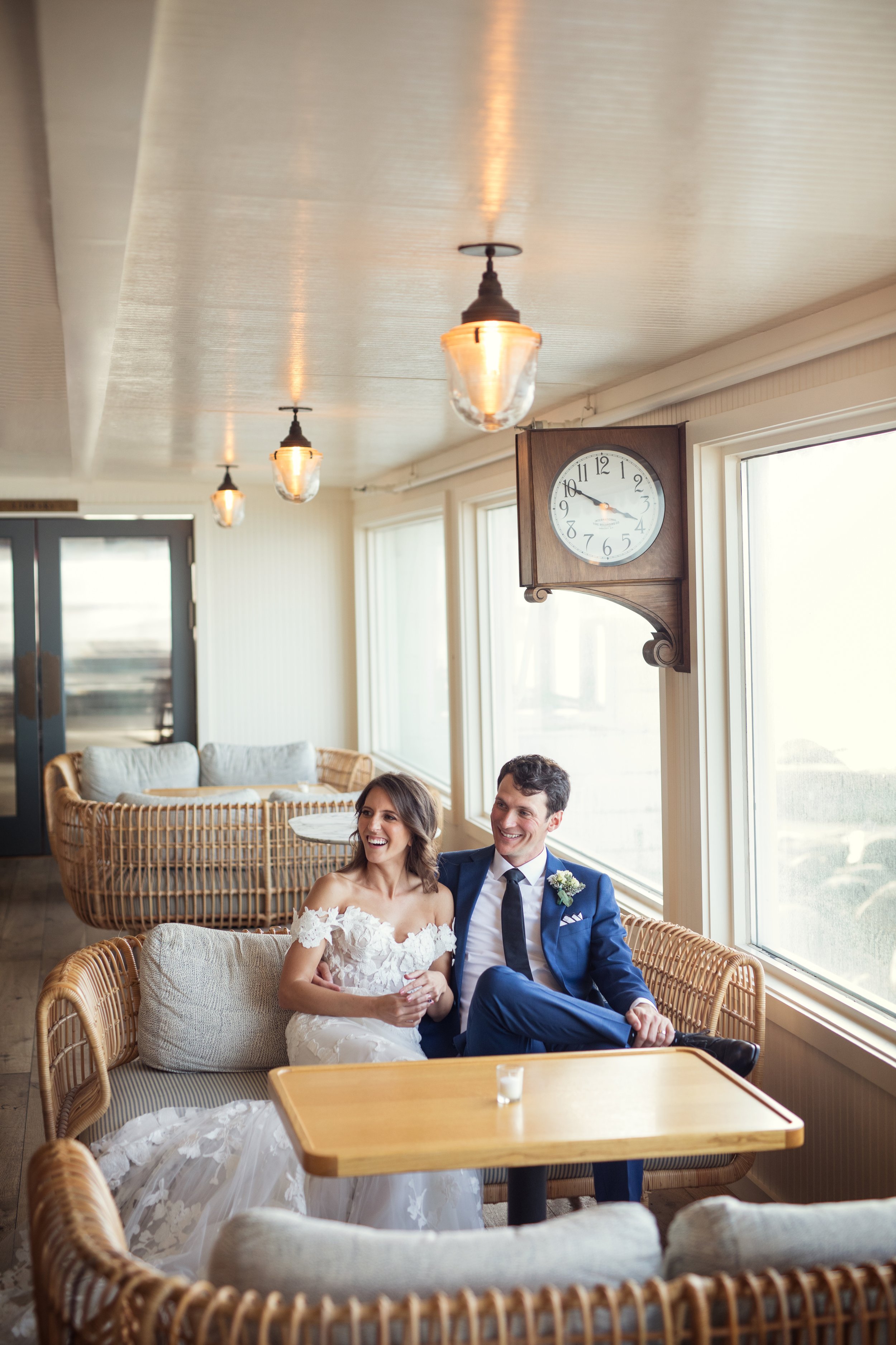 A bride and groom sitting together on a wicker sofa in a sunlit room, smiling at each other, with a clock on the wall showing 9:50 and lamps hanging from the ceiling.