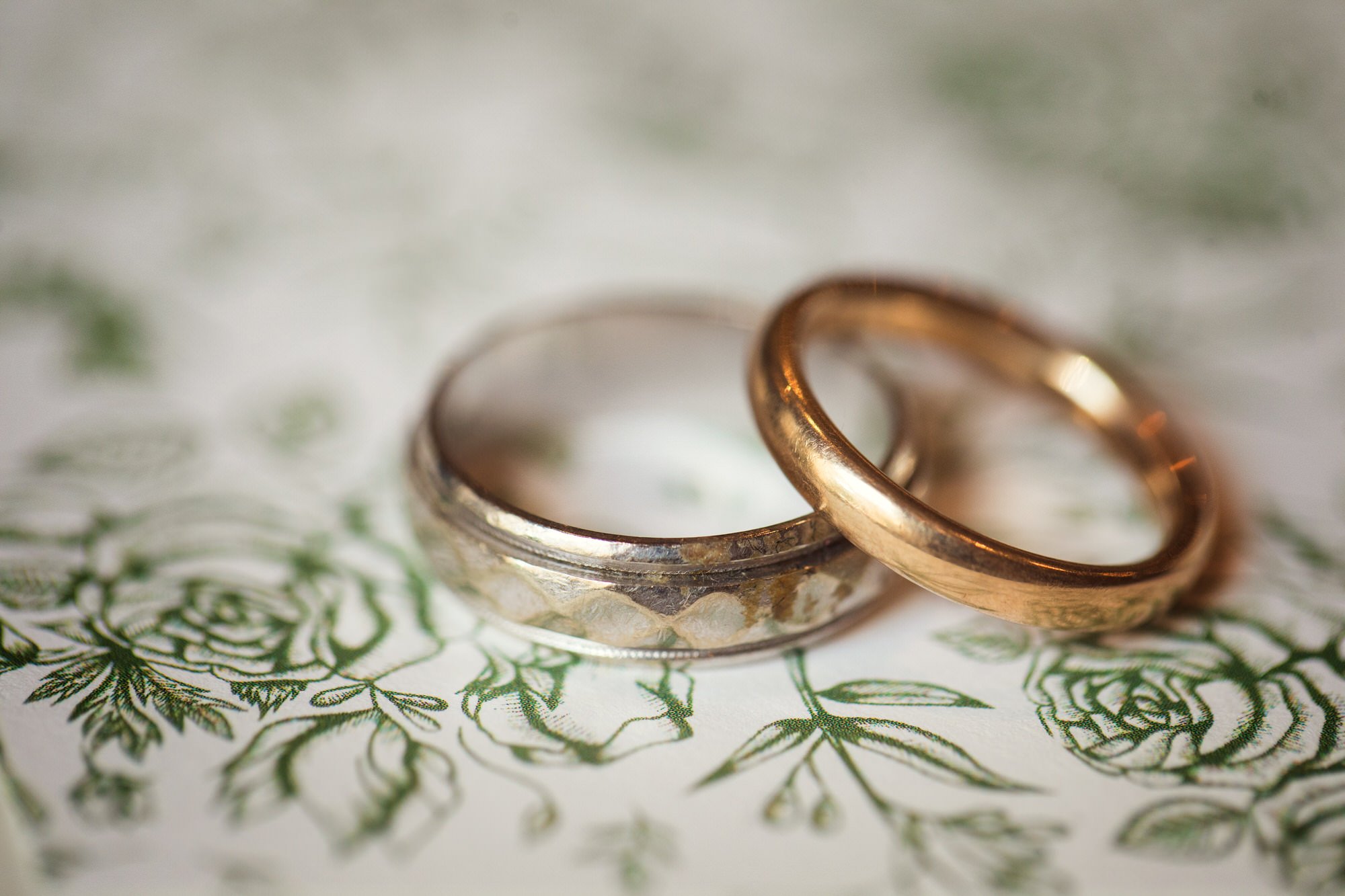 Close-up of two wedding rings, one silver and one gold, resting on a decorative green and white floral patterned paper.