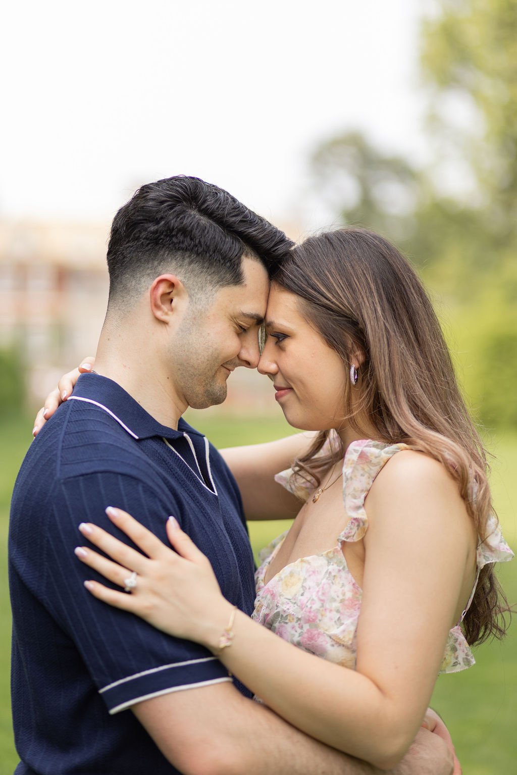 A couple embraces outdoors, forehead to forehead, smiling, with a green park and blurred trees in the background.