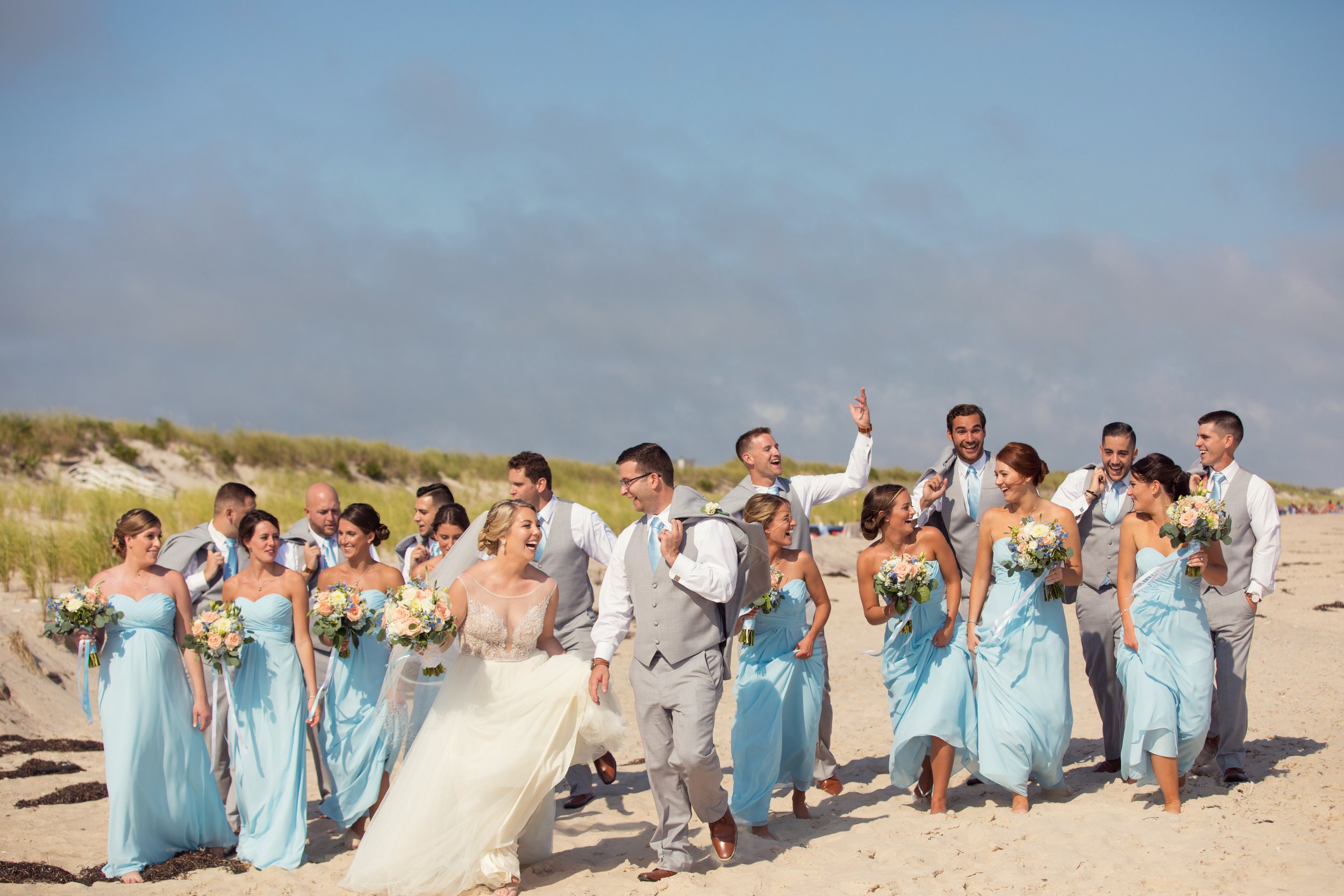 A wedding party walking along a beach, with bridesmaids in light blue dresses, groomsmen in gray vests and white shirts, and a bride in a white gown holding a bouquet, all smiling and laughing.