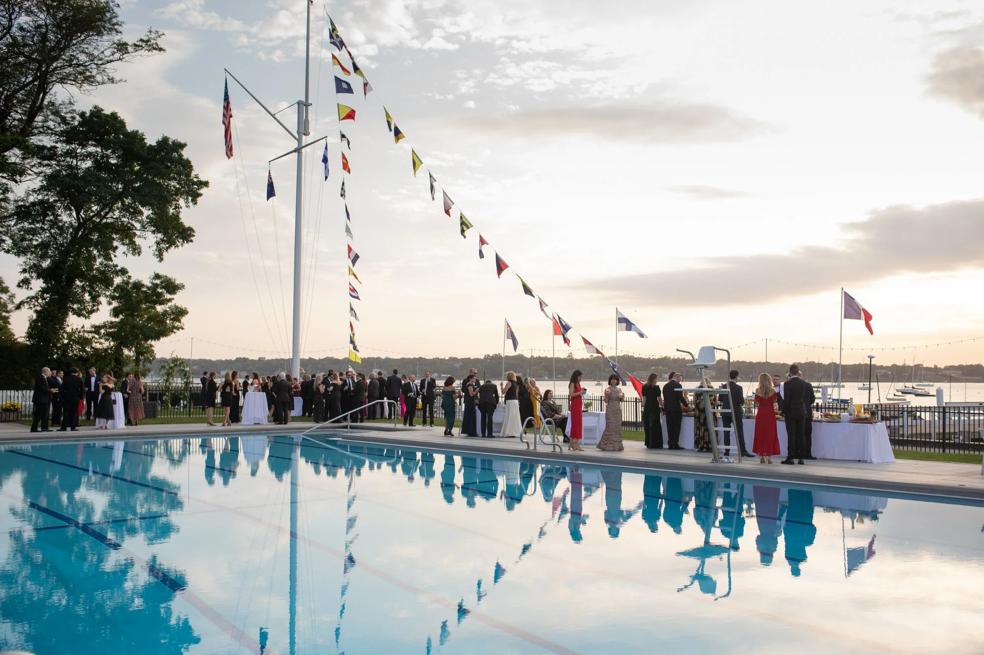 People at an outdoor party by a swimming pool near a waterfront, with flags and a sunset sky in the background.
