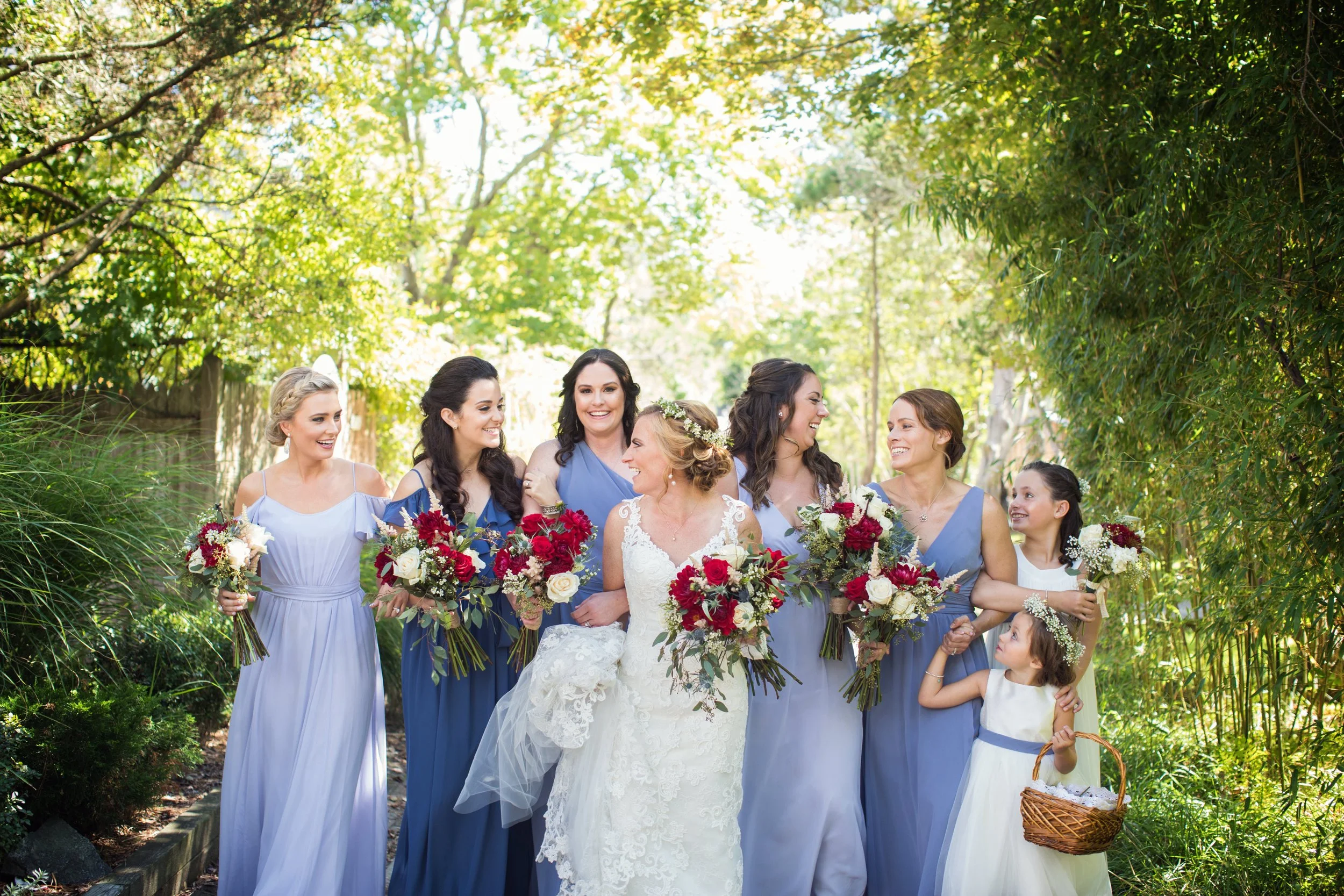 A bride and six bridesmaids standing outdoors in a lush green setting during a wedding celebration, all dressed in elegant dresses and holding bouquets of flowers.