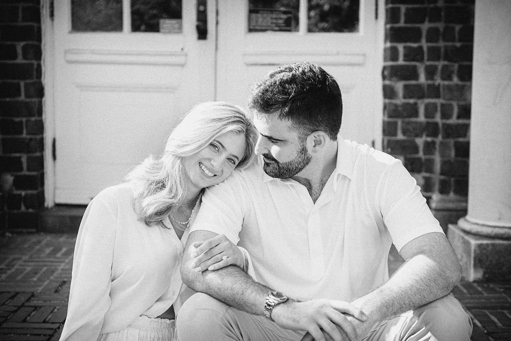 A woman and a man sitting close together outside, smiling and looking at each other, with a brick wall and a white door in the background.