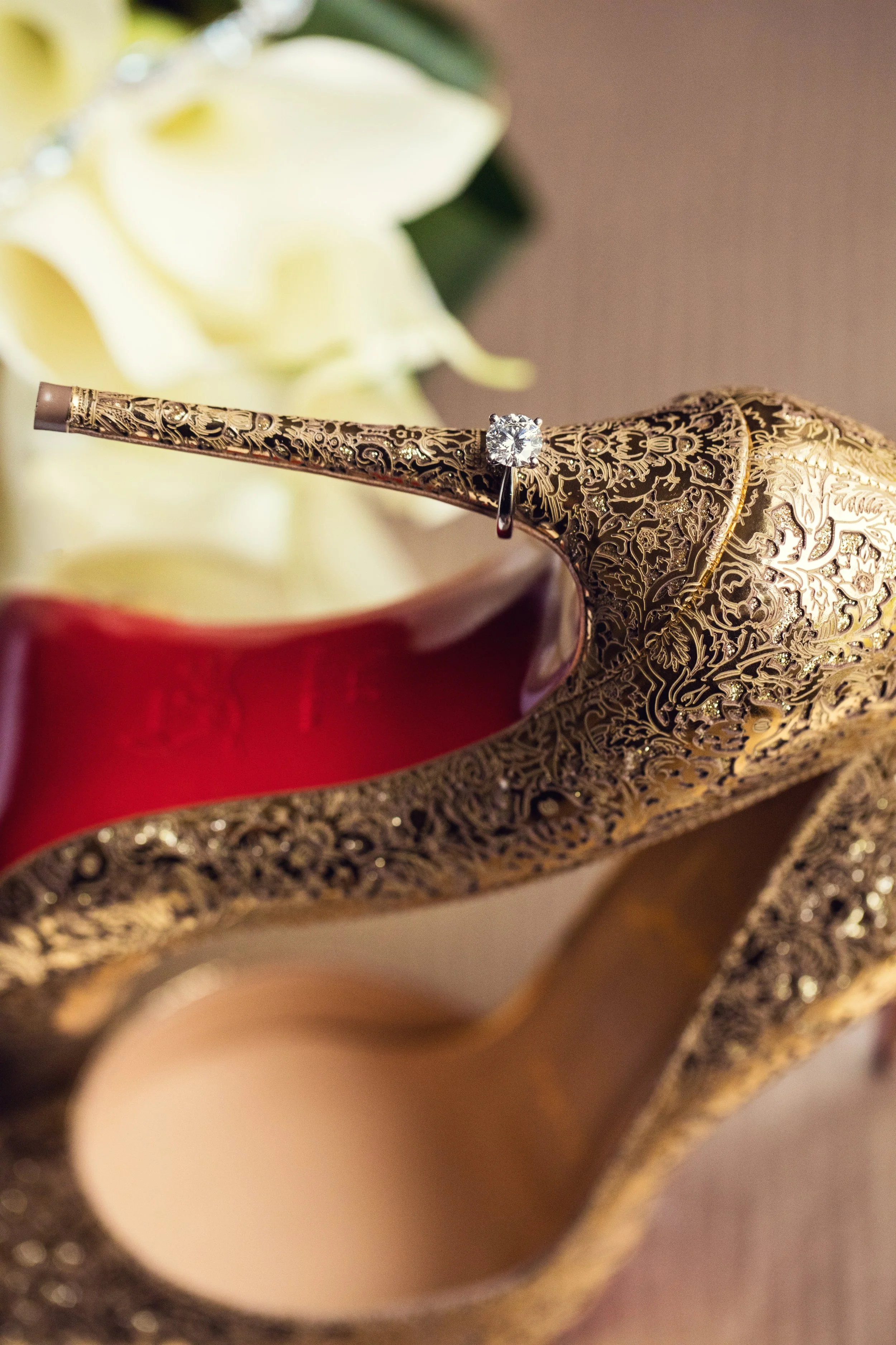 A close-up of a gold ornate high-heeled shoe with a diamond engagement ring placed on the heel, with a white flower in the background.