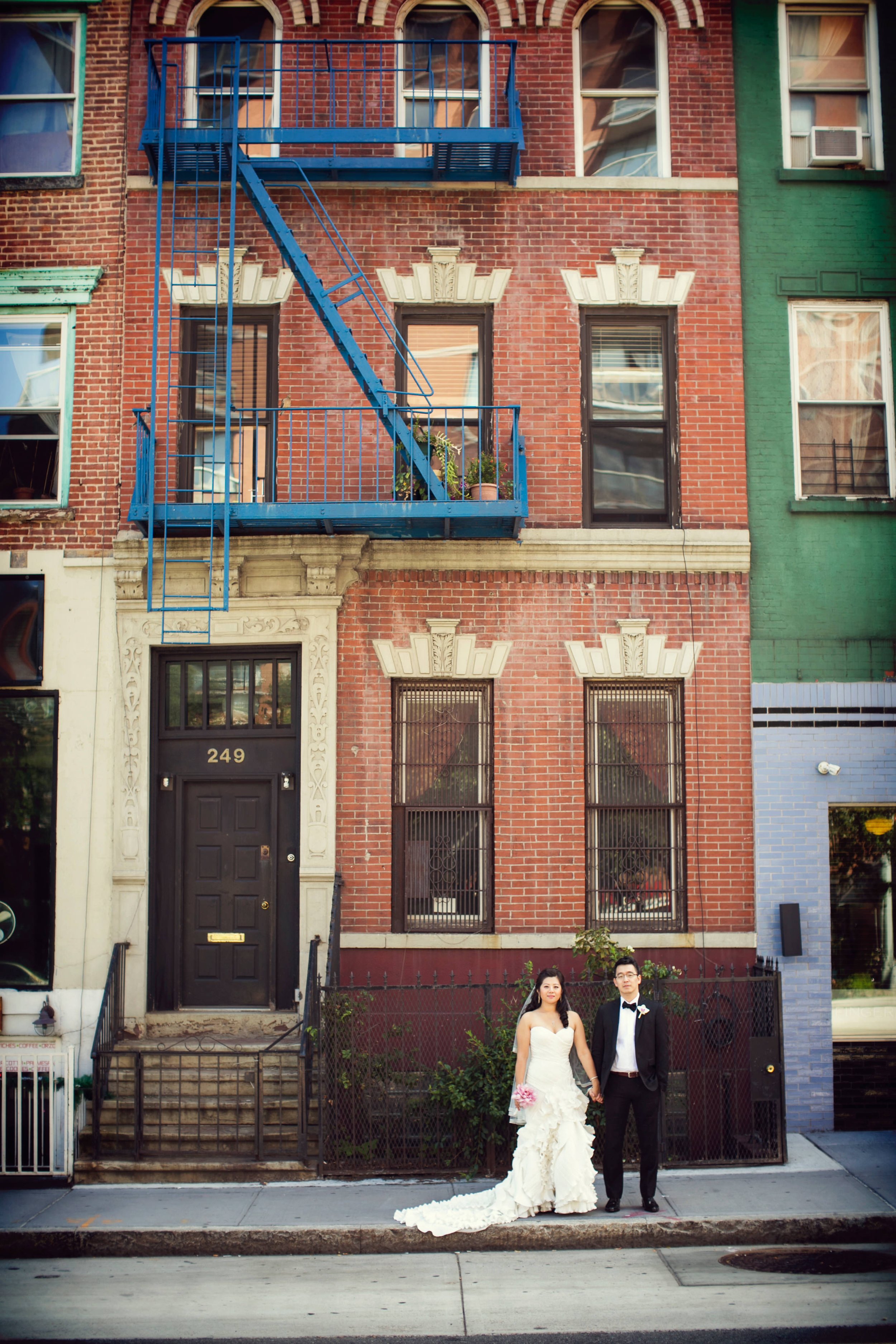 A bride and groom holding hands, standing in front of a red brick building with black door numbered 249. The bride wears a white wedding gown and holds a bouquet, while the groom wears a black tuxedo with a bow tie. The building has decorative window