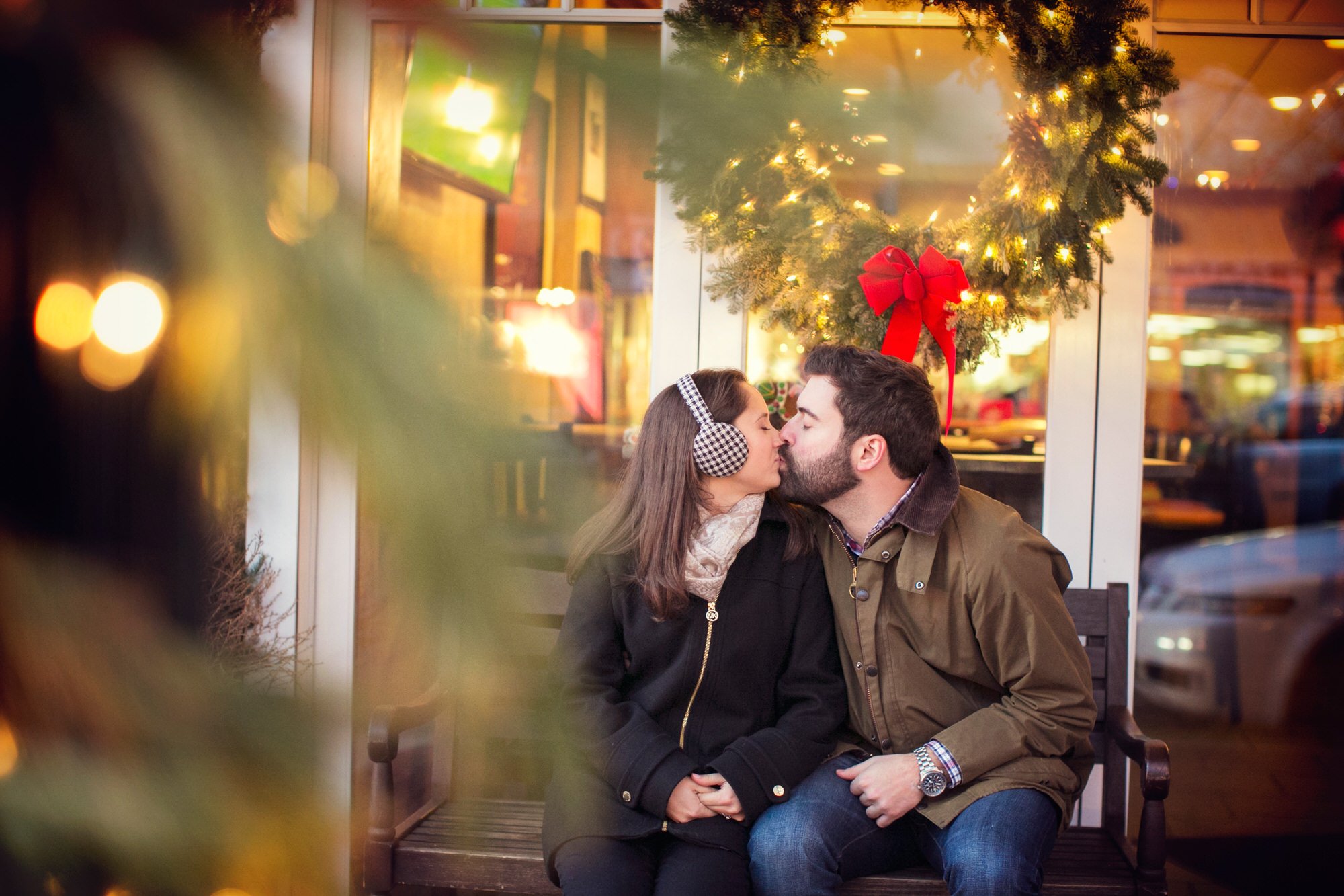 A couple sharing a kiss outside a cafe decorated with a Christmas wreath and red bow; blurred Christmas tree in the foreground.