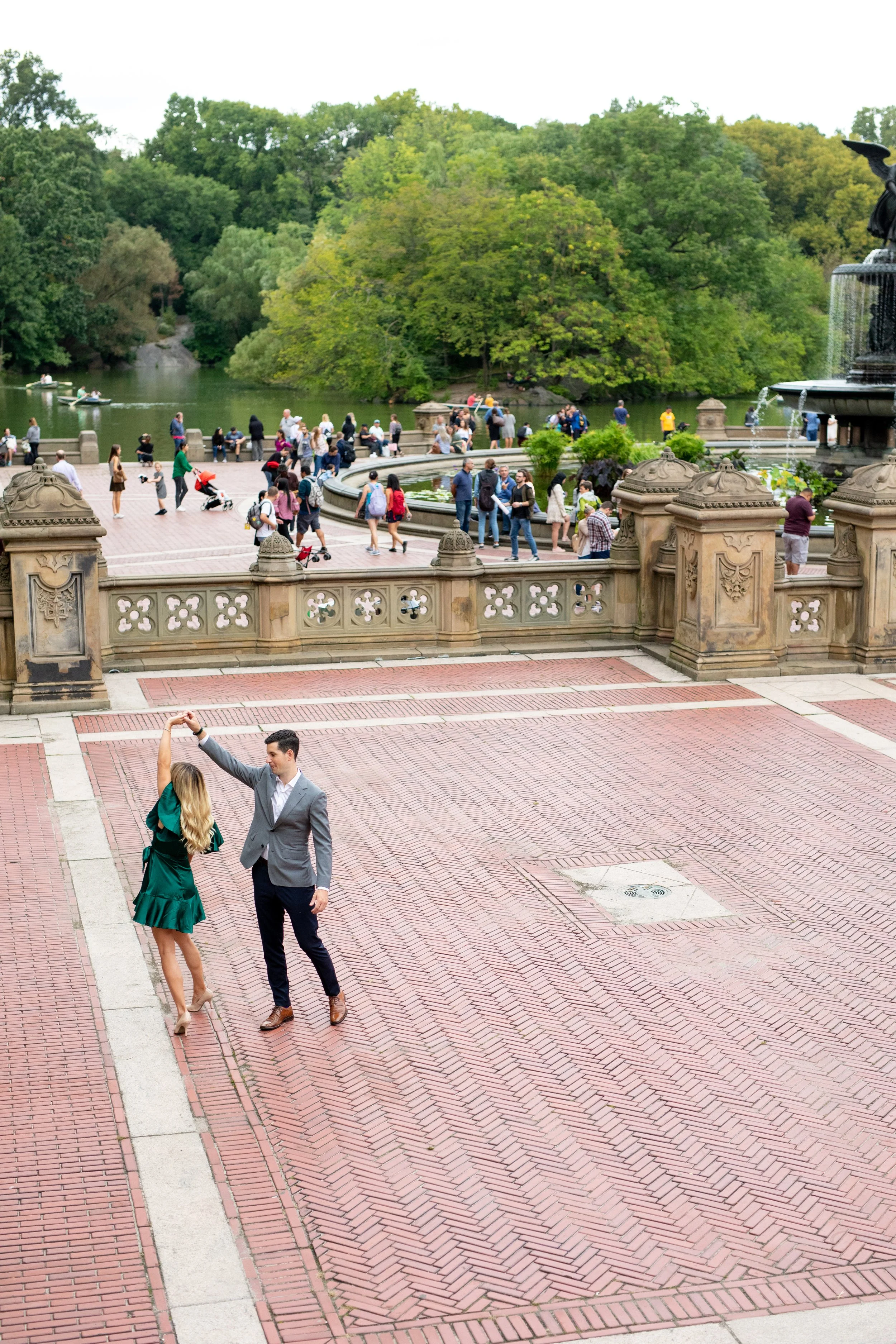 A young couple dancing on a brick patio near a fountain at a park, with many people in the background near a pond and lush green trees.