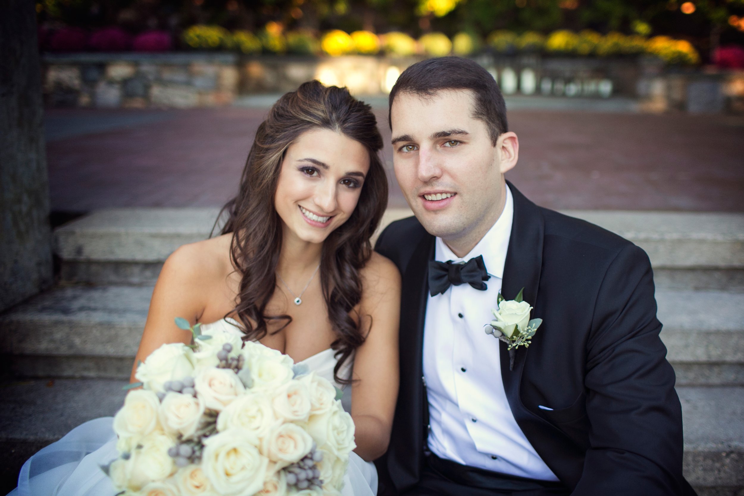 A newlywed couple sitting on a stone bench outdoors, smiling at the camera, with a bride holding a bouquet of white roses and a groom in a black tuxedo with a white shirt and black bow tie.