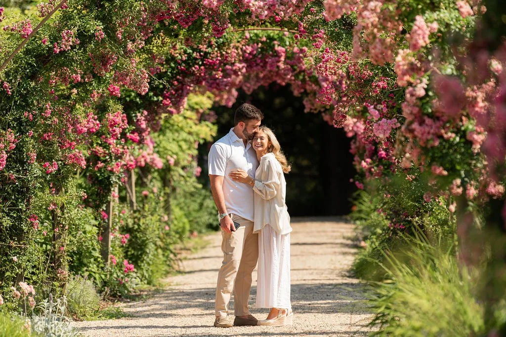 A man and woman standing close together under a floral archway, smiling and embracing in a garden.