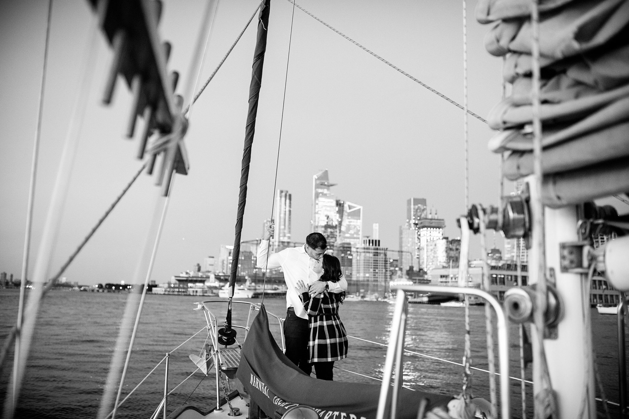 Black and white photo of a couple embracing on a sailboat with a city skyline in the background.