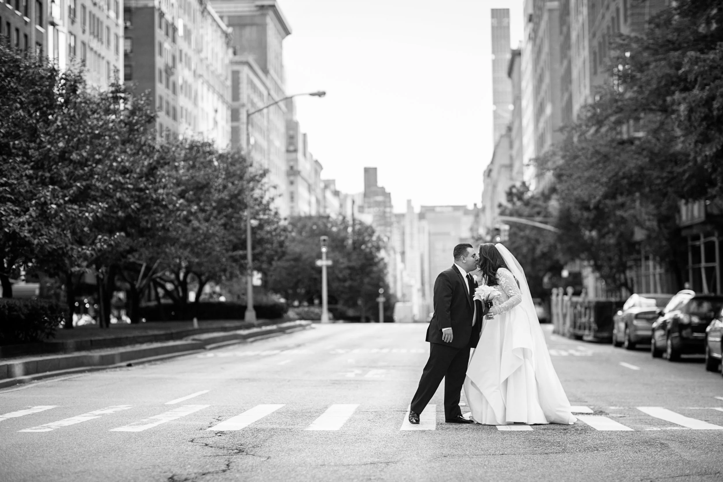 A black and white photo of a bride and groom kissing in the middle of an urban street with tall buildings, parked cars, and trees on either side.
