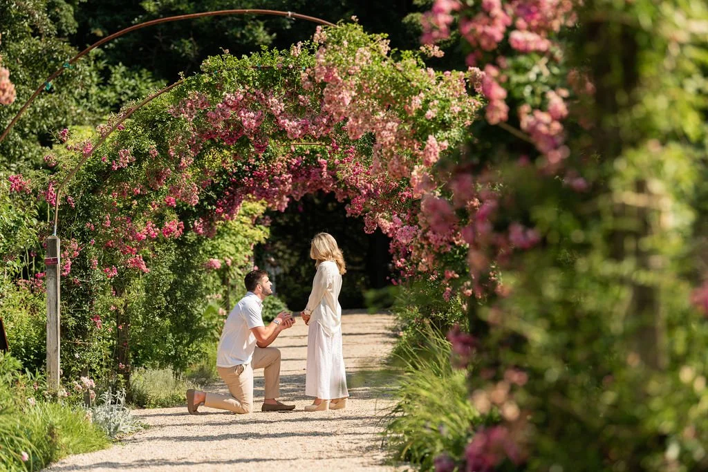 A man proposing to a woman in a garden with pink flowers and greenery.